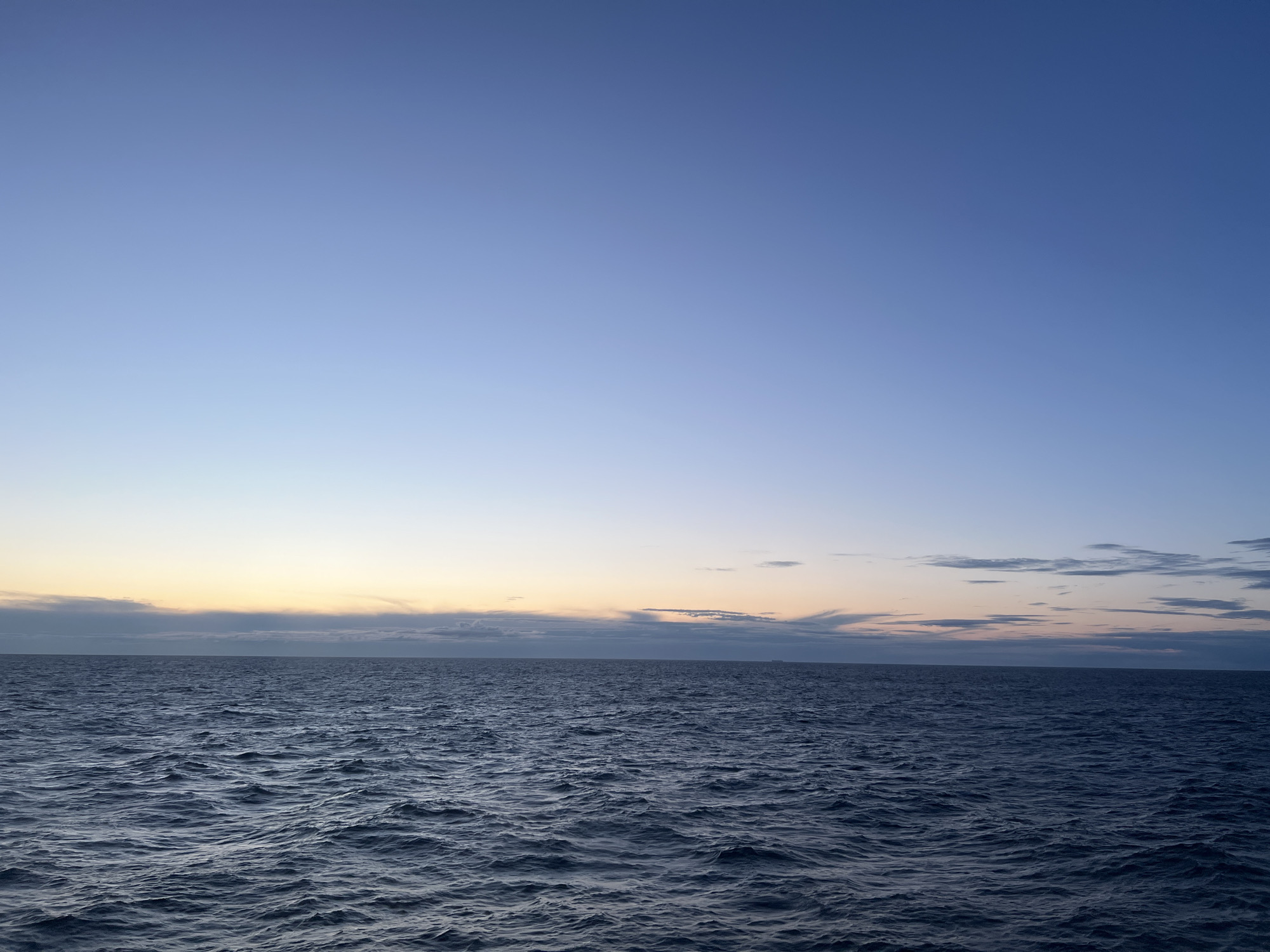 The image shows a wide expanse of dark blue water, rippling with small waves under a clear, pale blue sky. The horizon line is sharp and straight, dividing the water from the sky. Towards the horizon, a faint band of pale yellow and grey clouds is visible, suggesting either dawn or dusk. There are no people, animals, structures, or distinct landmarks in the frame. The scene is entirely natural, depicting an open body of water with a calm sky. The location is specified as Unknown, Poland, however, the image itself provides no visual clues to confirm this location.