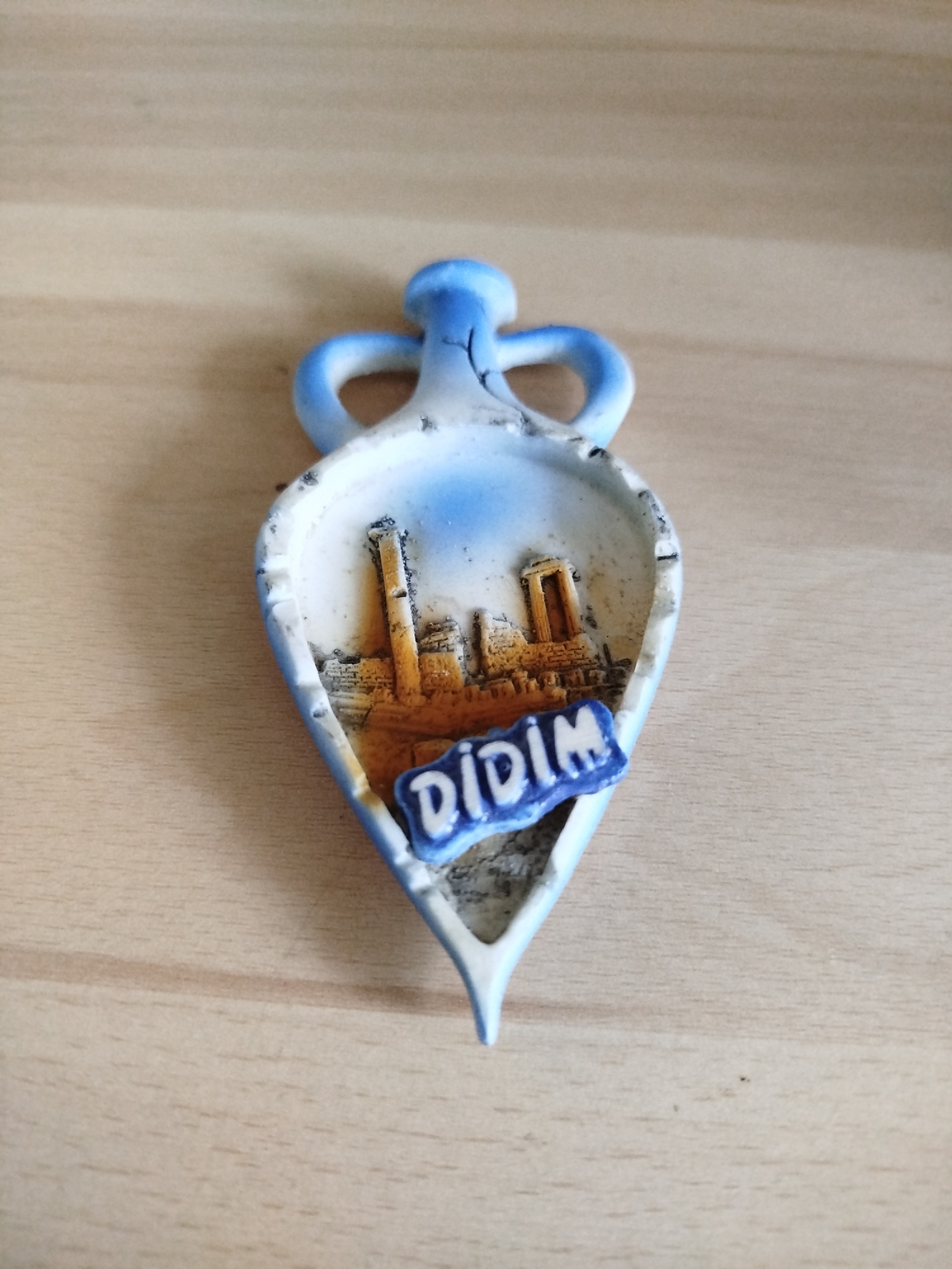 A souvenir featuring the ancient city of Didim, Turkey, shaped like a blue and white vase, is displayed on a wooden surface. The depiction within the vase shows building ruins. A blue plaque on the souvenir reads "DIDIM" in white letters.