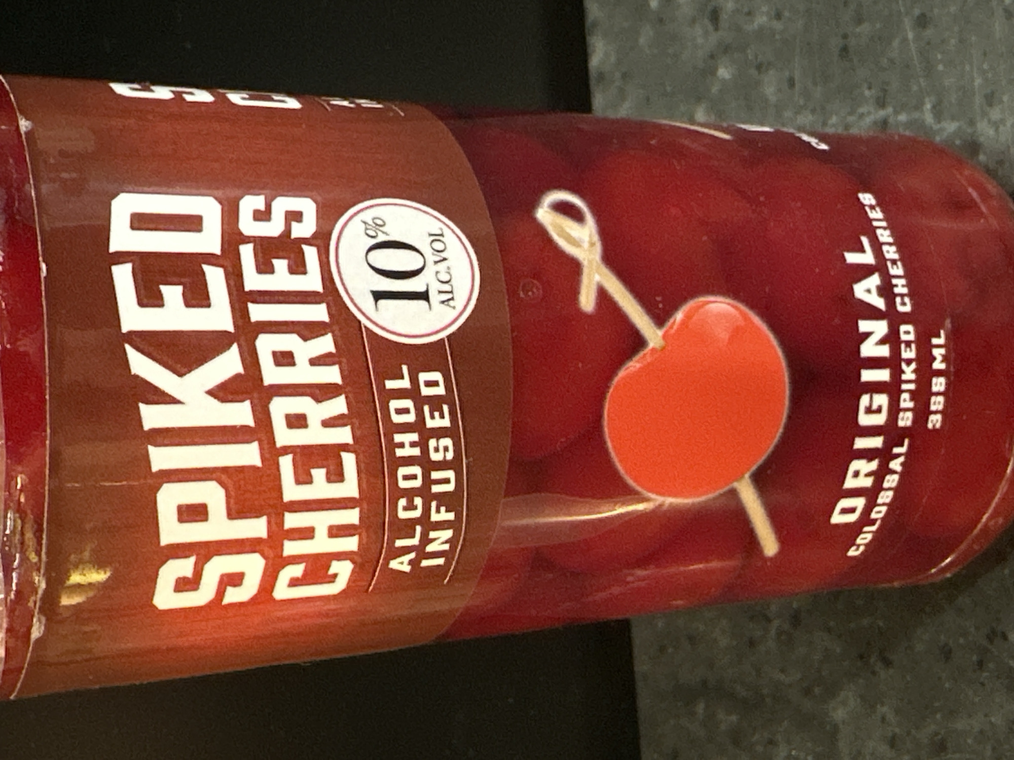 A close-up, eye-level shot shows the label of a glass jar filled with red cherries. The jar has a dark red label with white text. The text reads "SPIKED CHERRIES" in large, bold letters. Below this, in smaller text, are the words "ALCOHOL INFUSED" and "10% ALC.VOL." The jar contains numerous red cherries, with one cherry impaled on a wooden pick positioned in the center of the frame. Further text on the label reads "ORIGINAL COLOSSAL SPIKED CHERRIES 398 ML." The background is dark and out of focus, suggesting an indoor setting. The scene is located on a countertop in Camp Hill, United States of America.