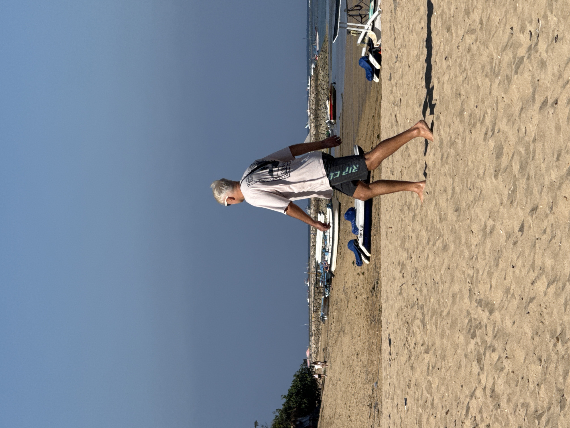 A man with gray hair and sunglasses is walking barefoot on a sandy beach in Denpasar, Indonesia. He is wearing a light-colored t-shirt with a graphic design on the back and dark shorts with the word "RIP CURL" printed on them. In the background, there are several boats moored in the water, a breakwater made of rocks, and some vegetation. The sky is a clear blue. The scene appears to be a typical day at a beach resort.