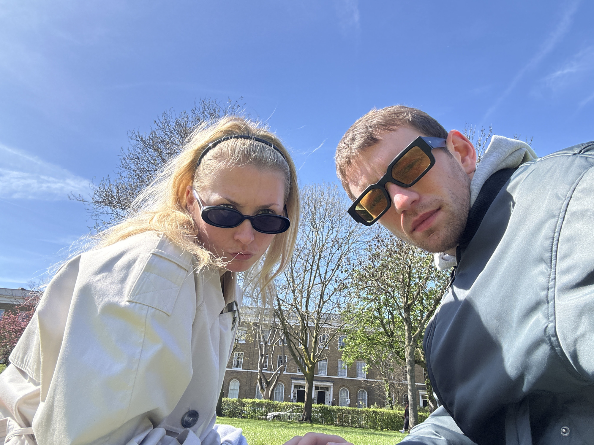 Two people, a woman and a man, are in the foreground, looking down at the camera. The woman has blonde hair pulled back with a black headband, is wearing sunglasses and a light-colored trench coat. The man has short brown hair, sunglasses and is wearing a grey hooded jacket. Behind them is a green lawn, trees, and a multi-story brick building with windows and doors. The sky is blue with some clouds. The location is likely in London, United Kingdom, potentially a park or garden area near historical buildings, given the architectural style of the building in the background.