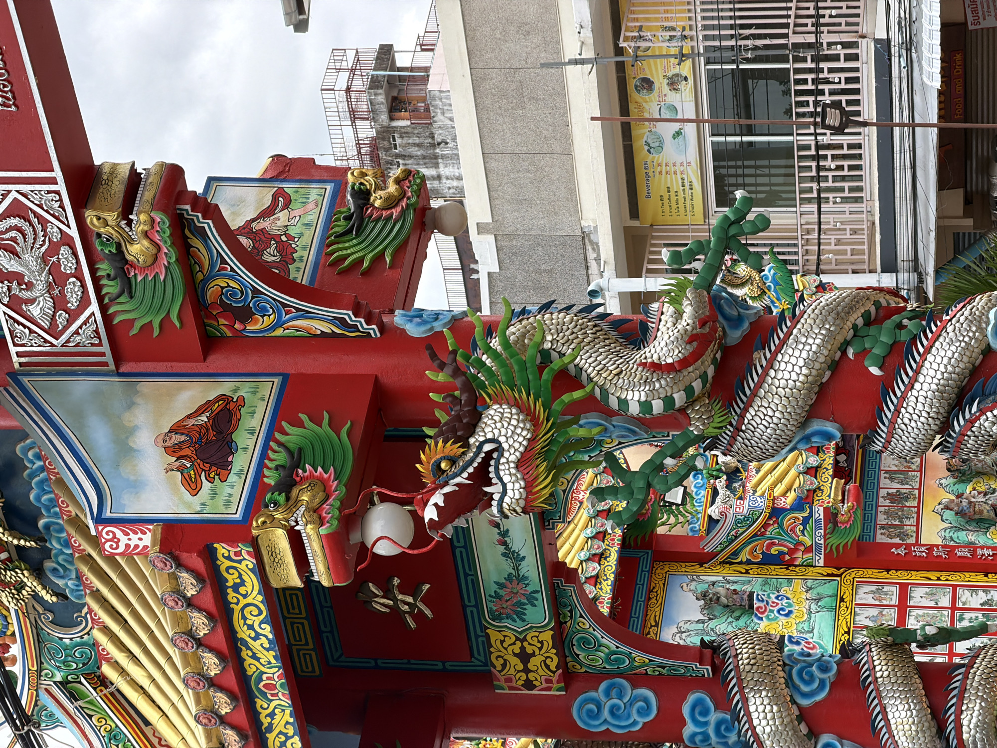 This media file is an upward-angled, close-up shot of the ornate, red-painted exterior of a temple or shrine structure, likely in Chiang Mai City Municipality, Thailand, given the context. The architecture is characterized by vibrant, traditional Chinese or Thai-Chinese motifs, featuring elaborate dragon sculptures, colorful paintings, and decorative accents.
Dominating the frame are detailed sculptures of serpents or dragons, with shimmering, scaled bodies, green, red, and yellow accents, and fierce expressions. These dragons are interwoven with the architectural elements, seemingly protecting or adorning the structure. Interspersed with the dragon sculptures are decorative paintings depicting figures in traditional attire. One painting shows a figure in orange robes walking across a green landscape, while another, partially visible, displays a grid of smaller scenes.
The structure itself is primarily a bright, bold red, with intricate gold and silver detailing. Elements like stylized waves, floral patterns, and what appear to be bamboo stalks are incorporated into the design. Small, decorative gargoyles or dragon heads with green manes are also visible. The sky above is overcast, suggesting a cloudy or possibly late afternoon/early morning time.
In the background, glimpses of a modern urban environment are visible, including buildings with balconies and windows, utility poles, and signage. One sign in particular, a yellow menu board with Thai text and images of food items, provides a clear indication of the location's context as a commercial area within Thailand. The overall impression is one of rich cultural artistry and traditional symbolism set against a backdrop of everyday urban life.