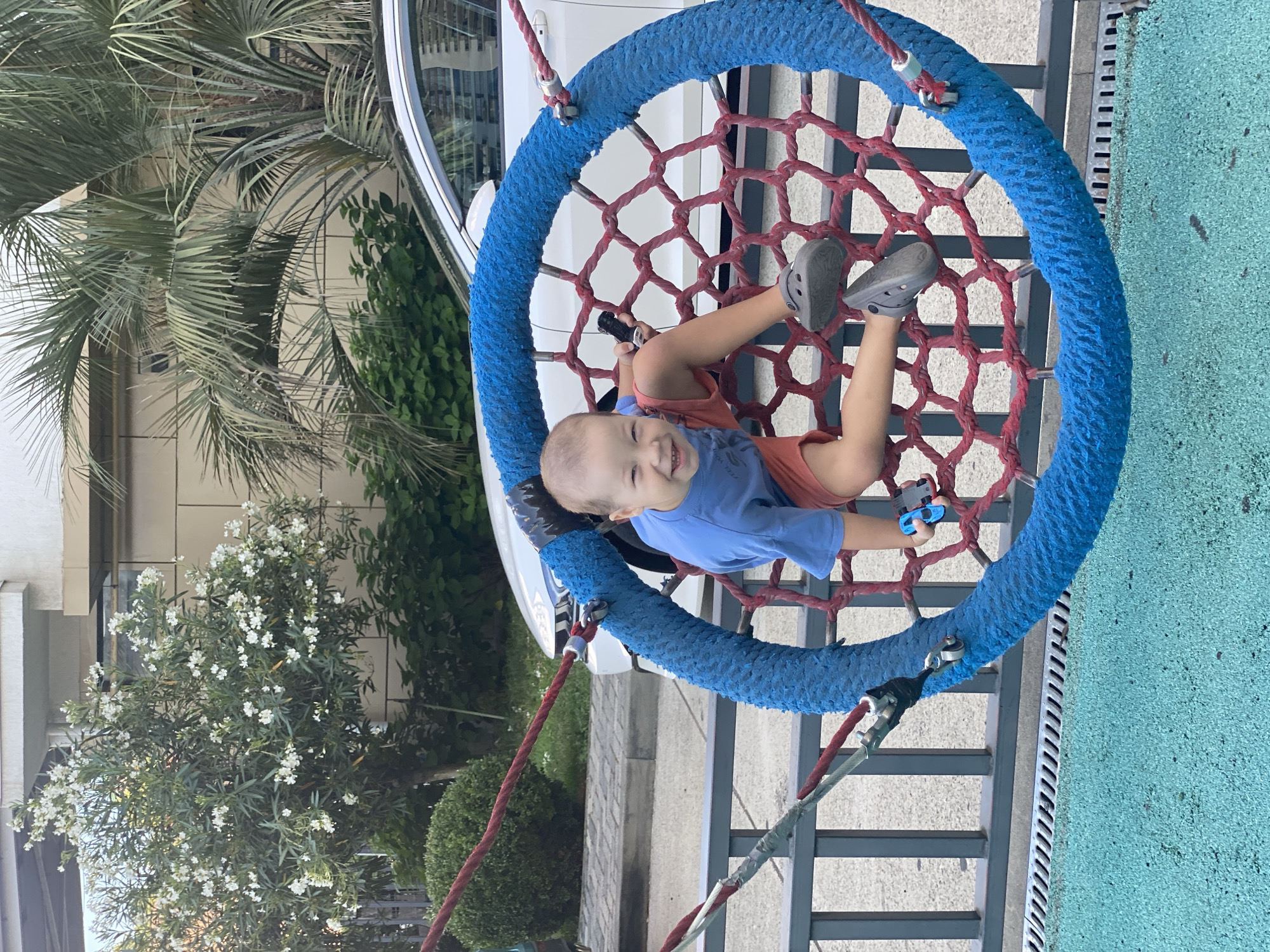 A young boy is playing on a circular swing at a playground. He is wearing a blue t-shirt and orange shorts, and grey Crocs. The swing has a blue rope rim and a red net interior. In his hands, he holds a small blue toy car. The playground has a blue and green rubberized surface. In the background, a white car is parked near a building with a beige facade. Palm trees and flowering bushes are visible, suggesting a warm climate. The fence of the playground separates it from a paved area. The overall scene appears to be a public park or playground in Sochi, Russia, given the presence of palm trees and the architectural style of the building.