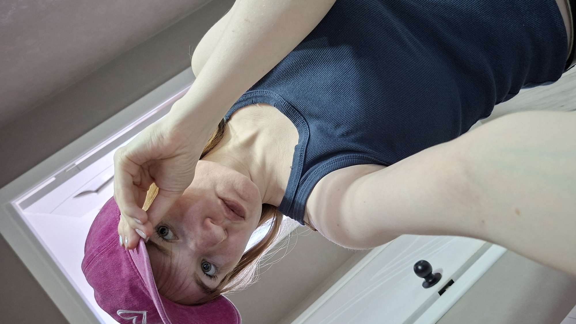 A woman wearing a navy blue tank top and a pink baseball cap is looking down at the camera. She is holding the brim of her cap with her right hand. The background includes a white door with a black doorknob, a light grey wall, and a white ceiling. The location appears to be indoors, possibly a home. There is no information available about the city of Chaikovsky, Russia, within this image.