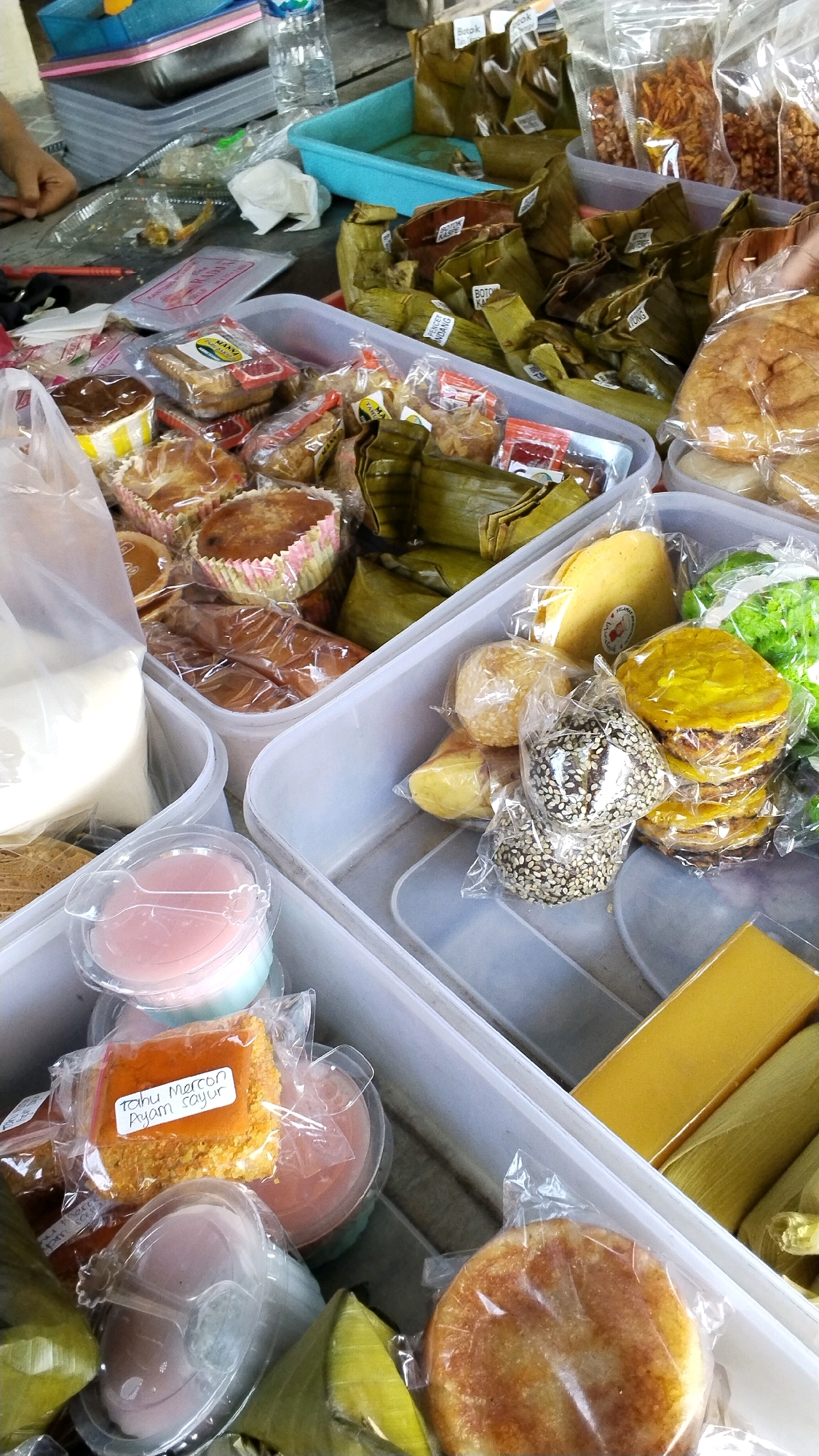 The image depicts a close-up, overhead view of a market stall displaying a variety of Indonesian food items, likely in Banjaragung, Indonesia. The scene is cluttered with numerous food packages, mostly wrapped in plastic or banana leaves, arranged in white plastic containers.
In the foreground, several items are visible: a round, golden-brown pastry in the bottom right, wrapped in plastic. To its left, a triangular, green item, possibly wrapped in banana leaves. Further left, a clear plastic container holding a pink and blue layered dessert. Next to that, a square item labeled "Tahu Mercon Ayam Sayur" (spicy tofu with chicken and vegetables), also in plastic. Several other small, individually wrapped food items are scattered throughout the containers.
In the middle ground, more food items are organized in plastic bins. To the left, there are small cakes or muffins in paper wrappers, some with a dark topping. To the right, a stack of yellow and green circular food items, possibly sweet cakes or pancakes, are visible, some dusted with sesame seeds. Behind these, several items wrapped in banana leaves are arranged.
In the background, more food items in plastic bags are seen, some appearing to be snacks like fried onions or crackers. A blue plastic tray contains more packaged food items wrapped in banana leaves. A water bottle and a portion of a person's hand and arm are visible on the left side of the frame, suggesting a vendor or customer interaction. The overall environment appears to be an outdoor market setting, with natural lighting.