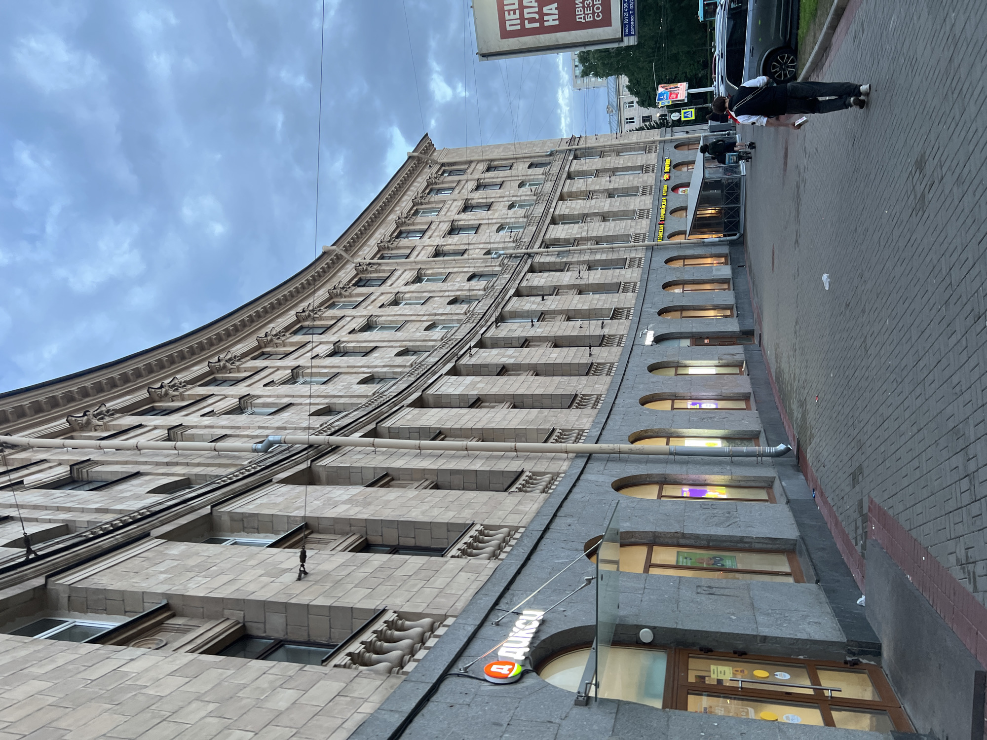 The image depicts a street scene in Saint Petersburg, Russia, in front of a large, curved, multi-story building with a stone facade. The building has many windows, some of which are illuminated, and architectural details like columns and balconies. A sign with Cyrillic lettering, reading "ДИКСИ" (DIXY), is visible on the lower level, indicating a store. The sidewalk is paved with brickwork and is bordered by a curb. A few individuals are visible walking along the sidewalk, including a person in a black vest and white shirt walking away from the camera, and another person on a scooter in the distance. Cars are parked along the street. The sky is overcast with clouds. A large billboard is visible on the right side of the image with Cyrillic text.