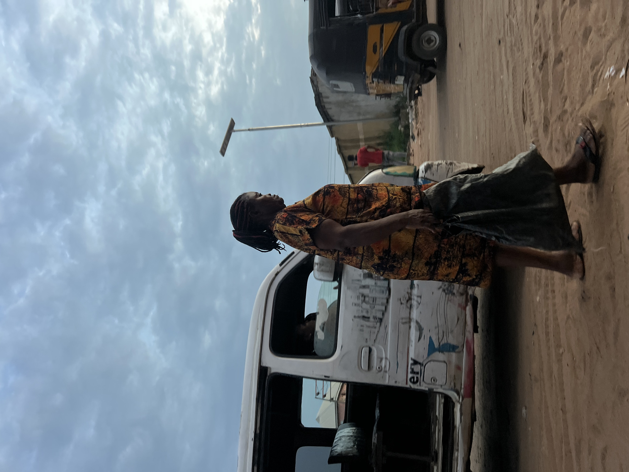 A woman with dark braided hair, wearing a patterned orange and yellow dress, walks along a dirt road. She carries a dark bag in her right hand. To her left, a white van is parked, with its door open. Inside the van, a person is visible in the driver's seat. A yellow and black auto rickshaw is to the right of the woman, and further in the background, a man in a red shirt stands near a wall. The sky is overcast with clouds. The scene is set in Nkpor, Nigeria.