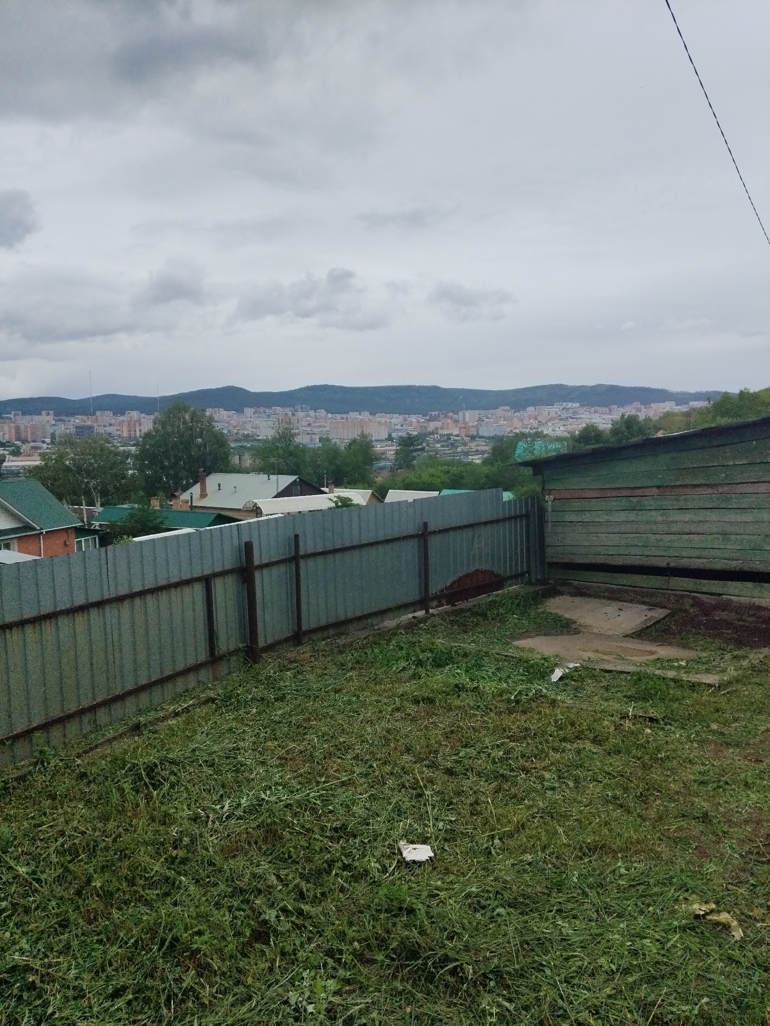 The image depicts a view from a hillside overlooking the city of Chita, Russia. In the foreground, there is a recently mowed lawn with piles of cut grass, a corrugated metal fence, and a dilapidated wooden structure on the right. Beyond the fence, several residential houses with pitched roofs are visible. In the background, the city of Chita stretches across the landscape, with numerous buildings of varying heights set against a backdrop of rolling, tree-covered hills. The sky is overcast with heavy grey clouds.