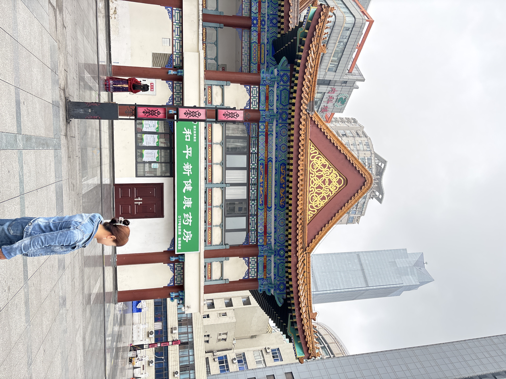 The image shows a street scene in Yuzhong District, Chongqing, China, with a traditional Chinese structure in the foreground. A woman wearing a blue denim jacket stands on the gray tiled pavement, looking down and to the left. To her right, a dark-colored lamppost is positioned next to a sign for "和平新健康药房" (He Ping Xin Jian Kang Yao Fang), a pharmacy. The traditional structure features colorful painted details and a roof with ornate gold patterns. Behind it, modern buildings and skyscrapers rise into the cloudy sky. Another person in a maroon outfit stands by a building in the background.