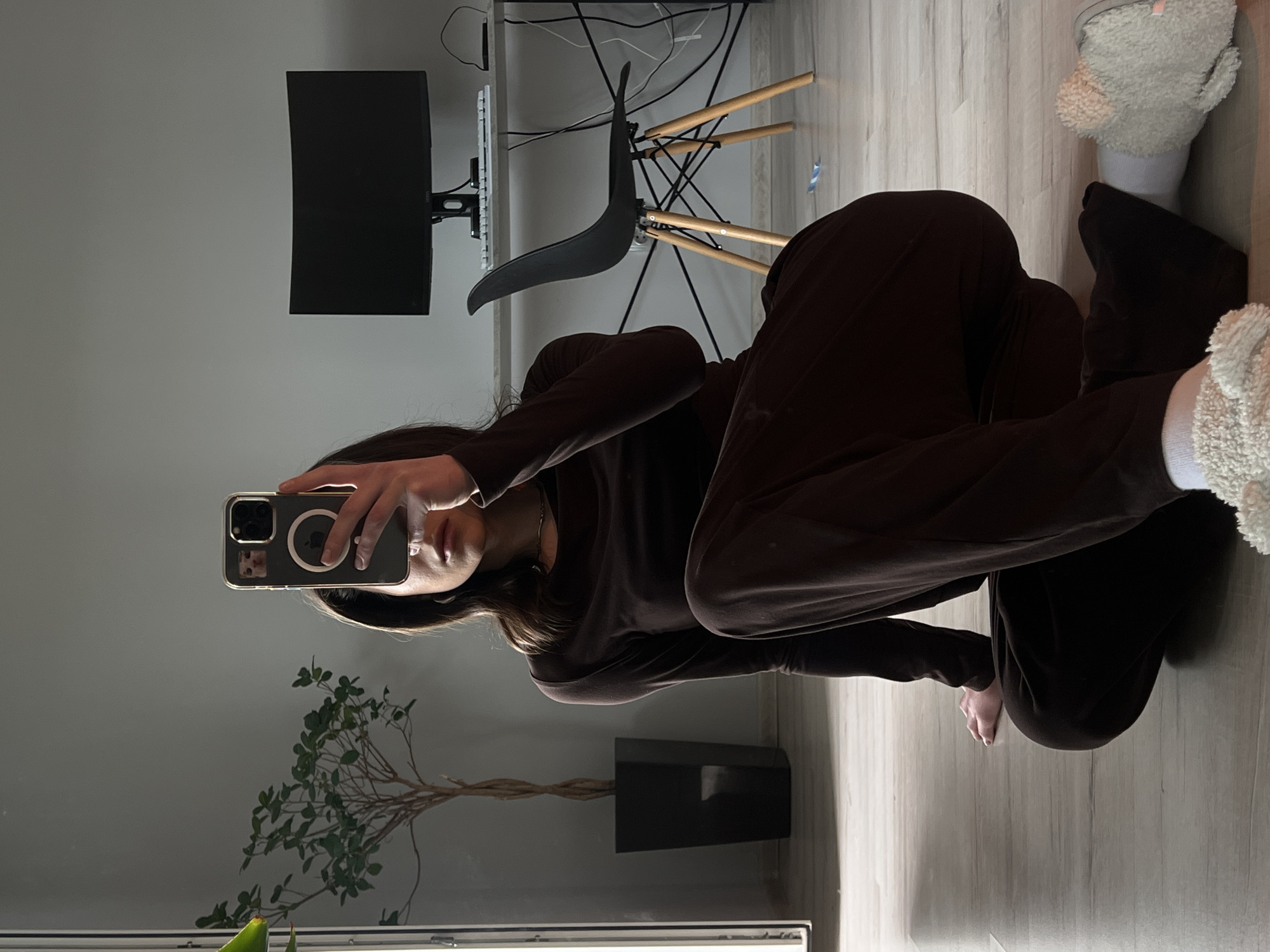 A person is sitting on a wooden floor in a room, taking a selfie in a mirror. The person is wearing a brown long-sleeved top and matching pants, with fluffy slippers on their feet. A plant in a dark pot is to the left of the frame, and a desk with a computer monitor and chair is in the background. The lighting suggests it is indoors, possibly in a residential setting. There is no discernible information that can associate this image with the city of Ternopil, Ukraine.
