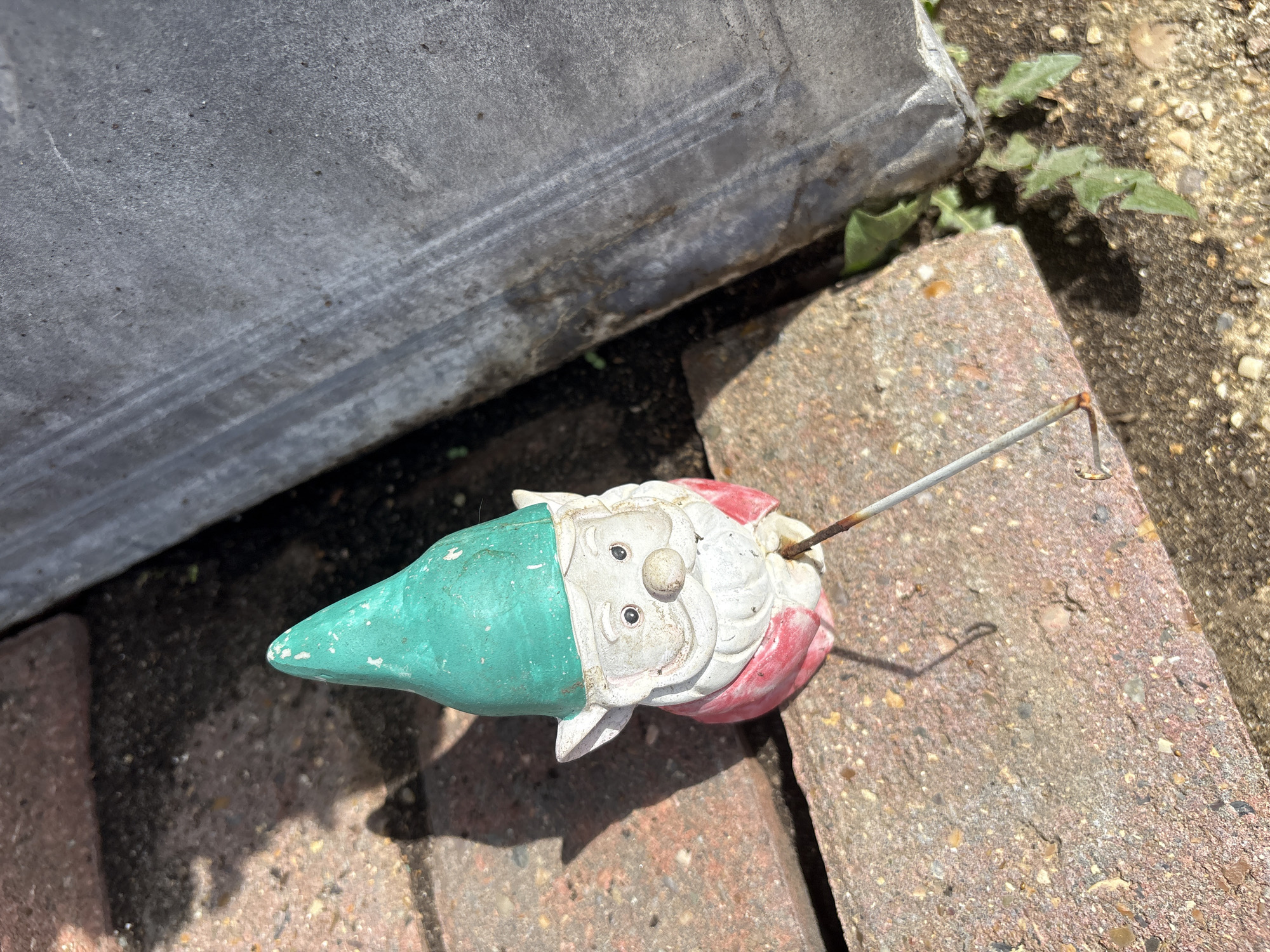 An overhead shot shows a small garden gnome lying on its side on a brick surface. The gnome is mostly white with a red coat and a green hat. Its face is visible, with two black eyes and a round nose. A thin metal rod extends from the gnome's back, bent at the end, and casts a shadow on the bricks. The background consists of dirt, some sparse green weeds, and a large, weathered gray object, possibly metal, that runs along the top left of the frame. The lighting suggests it is daytime, with sunlight casting distinct shadows. The scene appears to be outdoors, likely in a garden or yard.