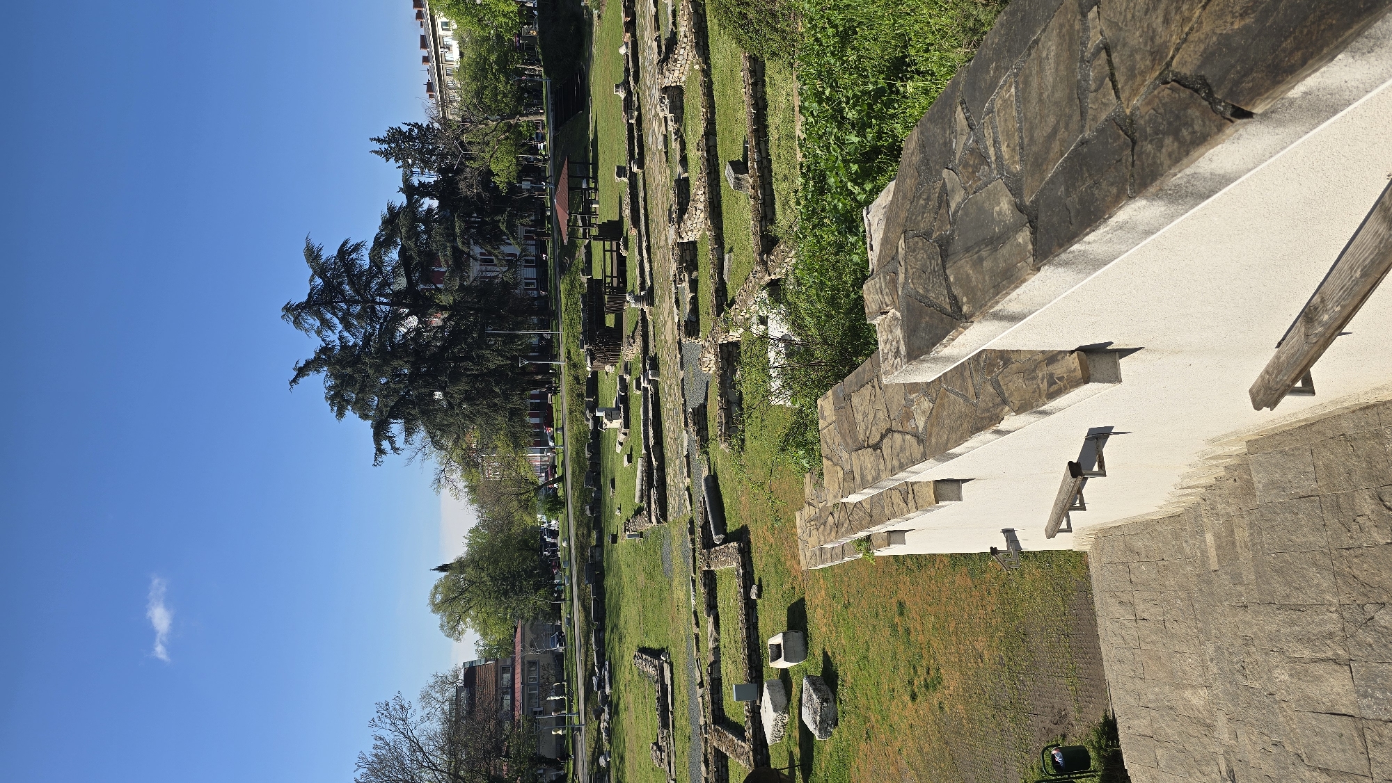This image depicts an archaeological park, identified as being in Stara Zagora, Bulgaria, under a clear, bright blue sky. The scene is bathed in strong daylight, casting distinct shadows and suggesting it's morning or early afternoon.
In the immediate foreground, to the right, are modern terraced structures built with light-colored concrete and clad with dark, irregularly shaped stone. These steps and seating areas feature integrated wooden benches. This contemporary architecture provides a viewing platform or seating for visitors to the site.
Beyond these modern elements lies the main archaeological area. Numerous stone foundations and remnants of ancient walls are visible, delineating the layouts of past buildings. These archaeological remains are interspersed within a well-maintained landscape of lush green grass. Scattered stone blocks and fragments are also visible throughout the grassy areas. In the mid-ground, a wooden structure, possibly a shelter or part of an exhibit, with a reddish-brown roof, stands amidst the ruins.
The background features a perimeter of tall, mature trees, including a prominent conifer with dense foliage. These trees partially obscure the urban environment, where glimpses of buildings can be seen, indicating the site is situated within or adjacent to a city.
The sky is a vibrant, clear blue, with only one small, wispy white cloud visible towards the left. There are no people visible in the image, nor is there any discernible text. The overall impression is one of historical tranquility and preservation within a modern urban setting.