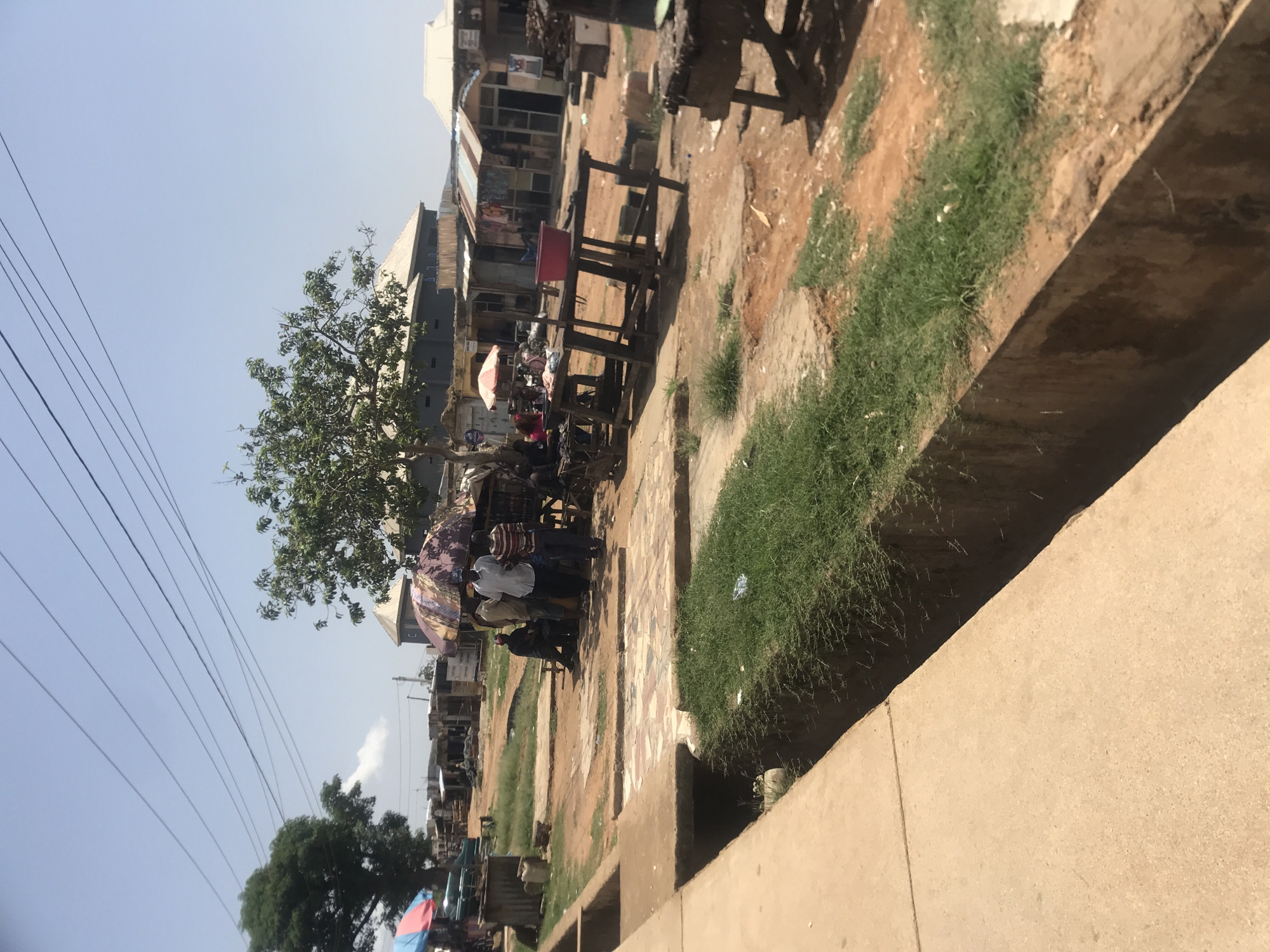 The image depicts an outdoor market scene in Rafin Par, Nigeria. In the foreground, a concrete walkway runs alongside a deep drainage ditch filled with grass and some debris. The middle ground features several wooden stalls and tables, some with umbrellas providing shade. A group of people, appearing to be vendors and customers, are gathered around these stalls.
In the background, more market stalls and simple structures with corrugated metal roofs are visible. A large tree with green foliage stands to the left, and another tree with sparser leaves is to the right. Power lines are strung across the clear blue sky. The ground is a mix of dirt and some paved areas with patterned tiles. The overall impression is a bustling, albeit somewhat rustic, local marketplace on a sunny day.
