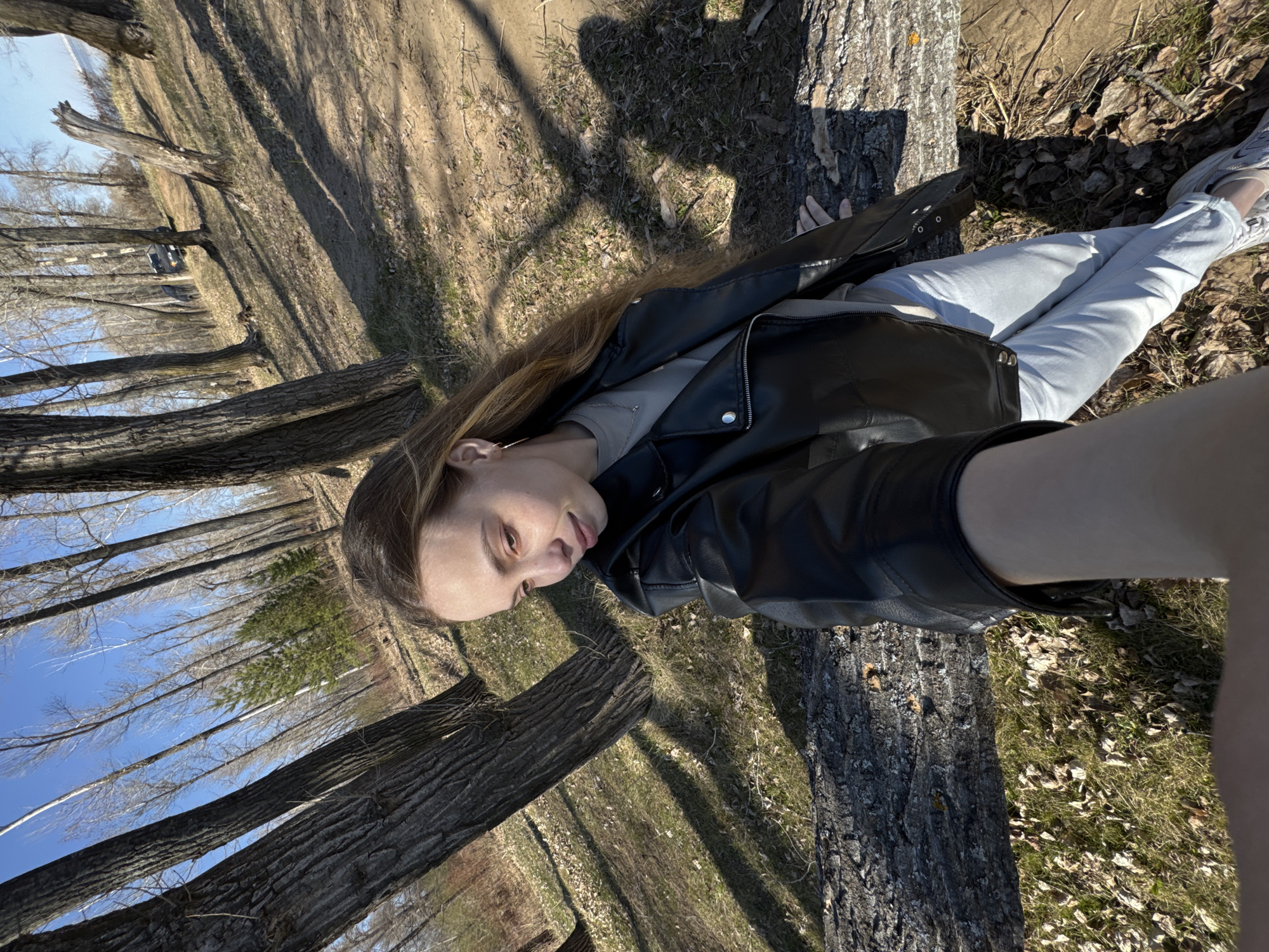 A young woman is photographed lying on a fallen tree in a wooded area. The woman is wearing a black leather jacket, a light grey t-shirt, and light blue jeans. She has long brown hair and is looking directly at the camera with a slight smile. The background shows a dense forest of bare trees and a blue sky. The location is in the city of Chaikovsky, Russia.
