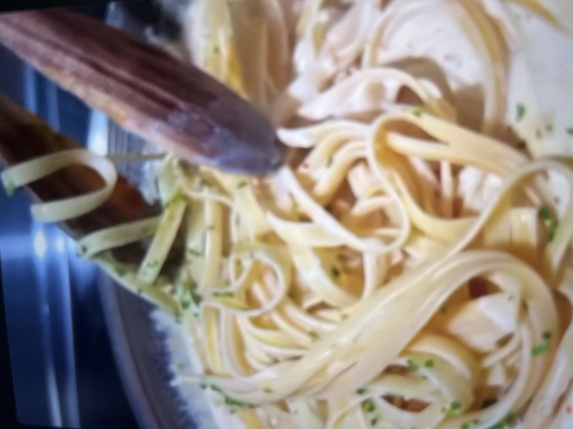The image shows a close-up view of fettuccine pasta being tossed with wooden tongs in a creamy sauce. Small green herbs are sprinkled over the pasta. The background is blurred and appears to be dark with some reflective surfaces, suggesting an indoor setting. There is no visible indication of the city of Abuja, Nigeria, or any specific environmental features that would place the scene there.