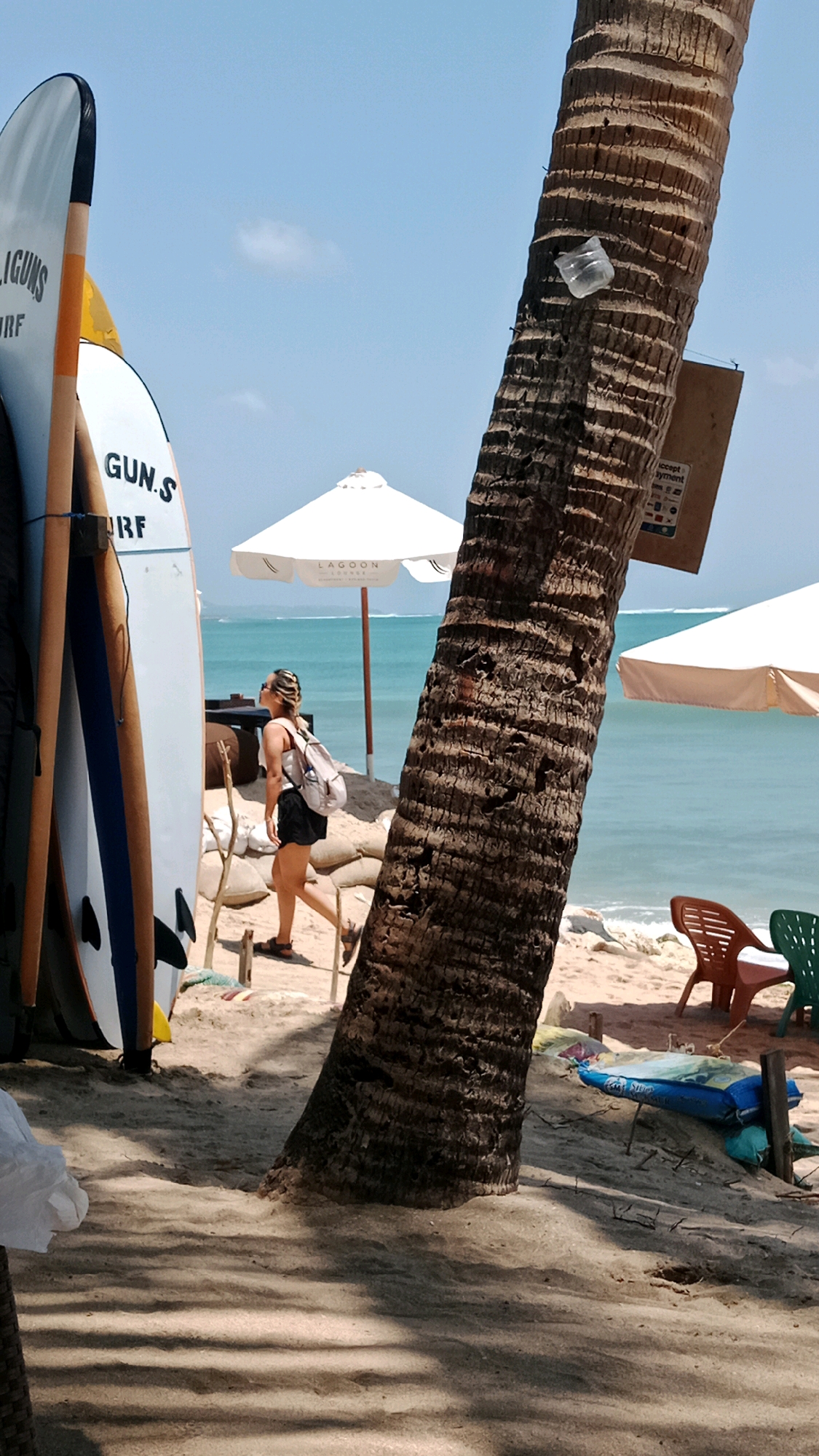 A full shot image shows a beachfront scene in Kuta, Indonesia. A tall palm tree with rough bark dominates the right side of the frame, its trunk angled towards the left. A piece of clear plastic is adhered to the trunk. The ground is covered in sand, with shadows of palm fronds creating patterns. To the left of the palm tree, several surfboards are propped upright. The surfboards are white with colorful accents and text that appears to read "GUN.S SURF". In the middle ground, a woman with her hair in a bun, wearing a white tank top and black shorts, walks to the left. She has a light-colored backpack on. Behind her, a white umbrella with "LAGOON LOUNGE" printed on it is set up. Beyond that, the turquoise ocean stretches to the horizon. In the background, another white umbrella is visible to the right, and a few chairs are on the sand. A blue inflatable object lies on the sand in the lower right.