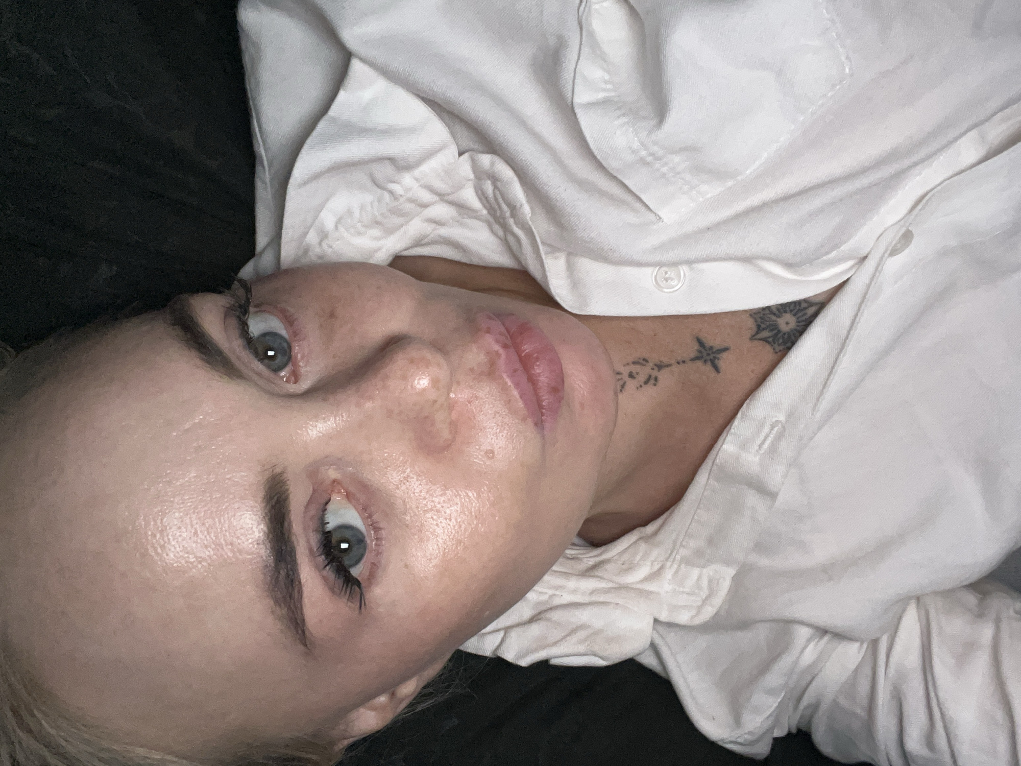 An overhead, close-up, selfie-style shot shows a woman lying down, likely in bed, with her head tilted and slightly propped up on her left side. Her blonde hair is pulled back from her face, and she is wearing a white collared shirt, the top button undone. She has light blue eyes, and her lips are slightly parted in a pout. There are freckles on her face, and she is wearing makeup, including mascara and possibly lip gloss. A tattoo of a star on a string and a floral design are visible on her neck and upper chest, just below the collar of her shirt. The background is dark and indistinct.