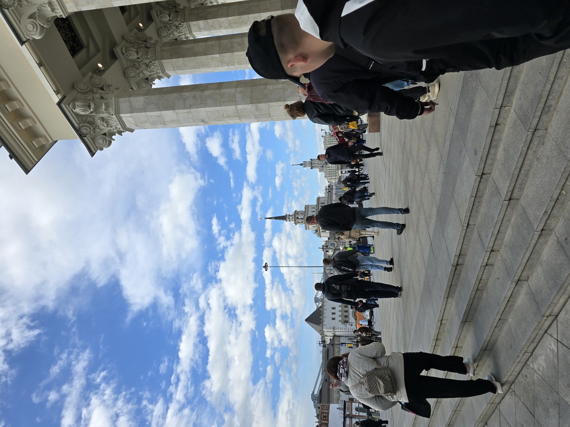 The image shows a view in Moscow, Russia. The sky is blue with white clouds. Several people are walking, standing, and sitting in the foreground. A building with tall, decorative columns is partially visible at the top. In the background, there are various buildings, including one with a pointed spire. The foreground features gray stone steps.