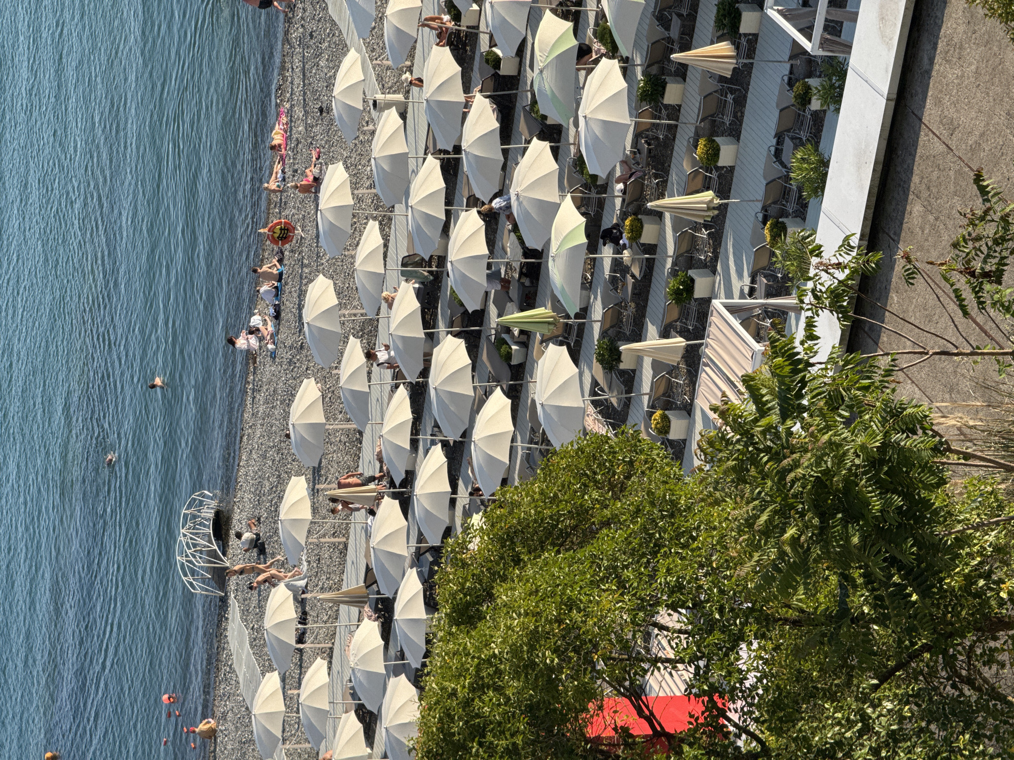 This image depicts a crowded beach scene at a resort, likely in Sochi, Russia, given the style of the umbrellas and the presence of a pebble beach. The beach is lined with numerous white and beige umbrellas, providing shade for lounge chairs arranged in neat rows. People are scattered across the beach, some sunbathing on towels, others swimming in the turquoise water, and some walking along the shoreline. A small white arched structure, possibly a part of a pier or art installation, is situated at the water's edge. Lush green trees and vegetation frame the lower part of the image, adding a natural element to the man-made resort setup. The overall impression is one of a busy, sunny day at a popular seaside destination.