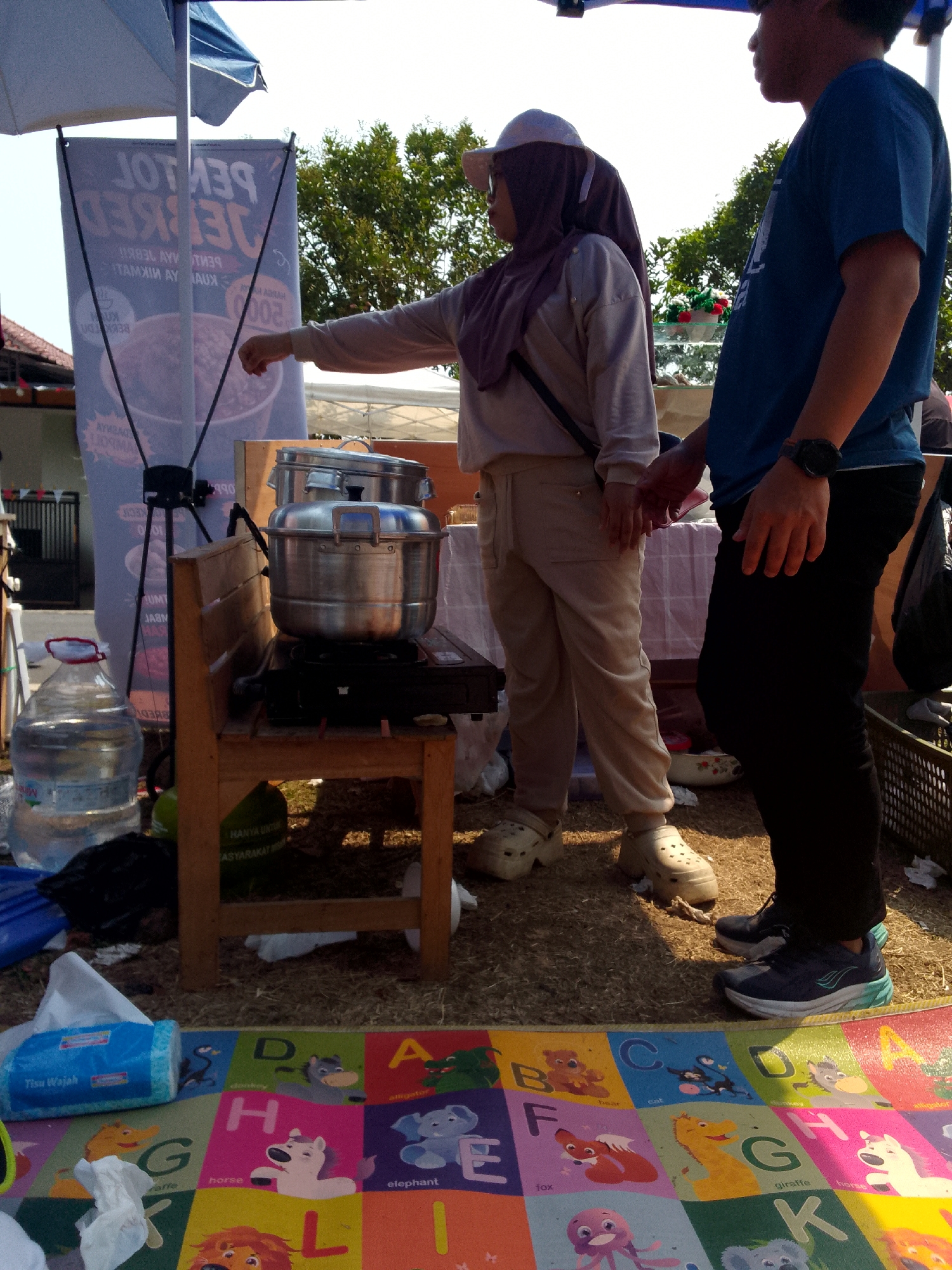 Two individuals, a woman wearing a purple hijab and a light-colored outfit, and a man in a blue t-shirt and black pants, stand in front of a food stall at what appears to be a street market or outdoor event. The woman is pointing towards a steaming pot on a portable stove. Behind them, a large banner with Indonesian text is visible, advertising "PENTOL DECK". The environment suggests a warm day, with a blue umbrella providing shade overhead and trees in the background. The ground is covered with straw or dry grass, and an alphabet mat with various animals is spread out in the foreground. A large bottle of drinking water and a pack of facial tissues are also present. The scene is located in Mandalawangi, Indonesia.