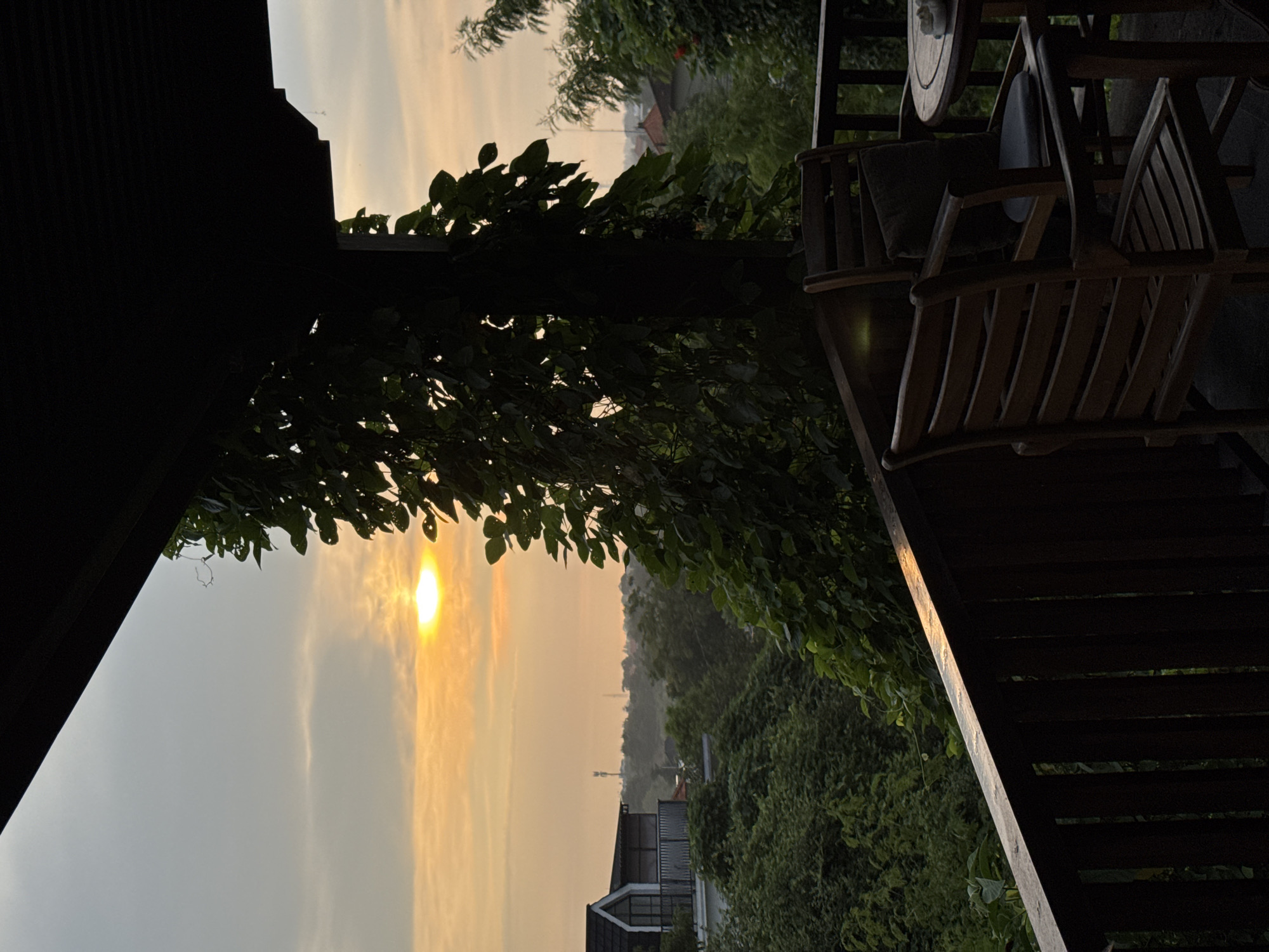 Here is a description of the image:
The image depicts an outdoor scene, possibly a balcony, at sunset or sunrise in Ungasan, Indonesia. The warm glow of the sun is visible through foliage. In the foreground are wooden chairs and a small table, part of the balcony structure. Beyond the balcony, the landscape is filled with lush greenery. A building with a corrugated metal roof is partially visible. In the distance, a communication tower can be seen rising above the trees.