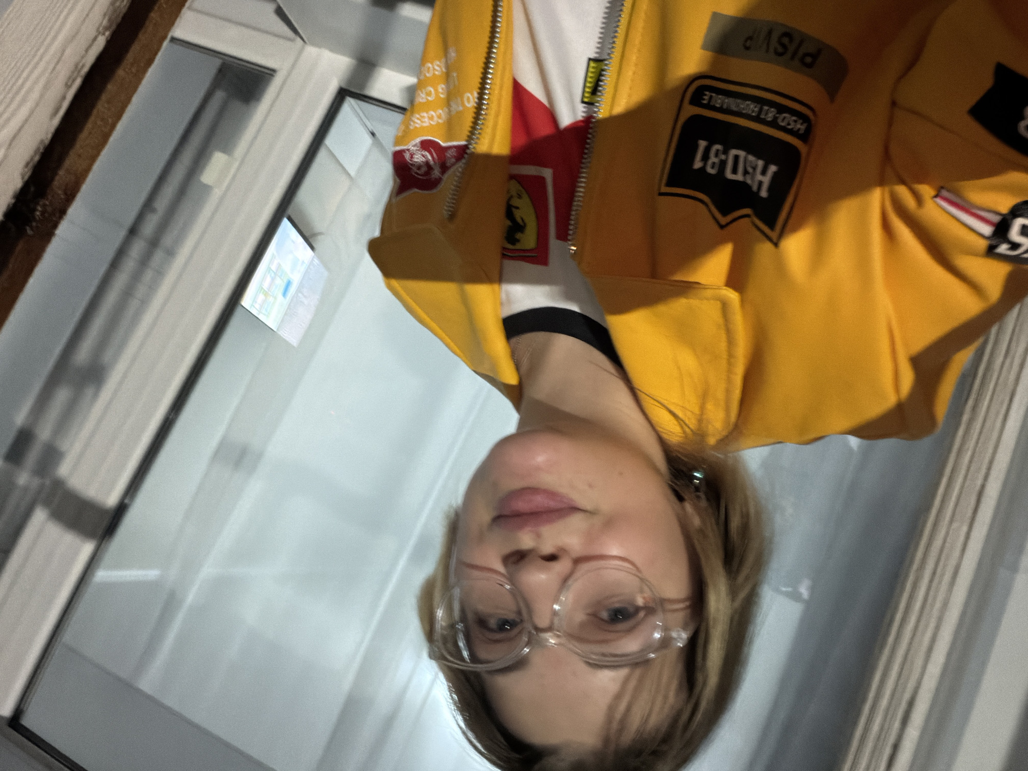 A young woman in a yellow jacket with racing-themed patches and logos (including a Ferrari logo) is photographed indoors. She is wearing clear-framed eyeglasses. The background appears to show a window and part of a wall. There is no verifiable evidence to establish the location as Kazan, Russia.