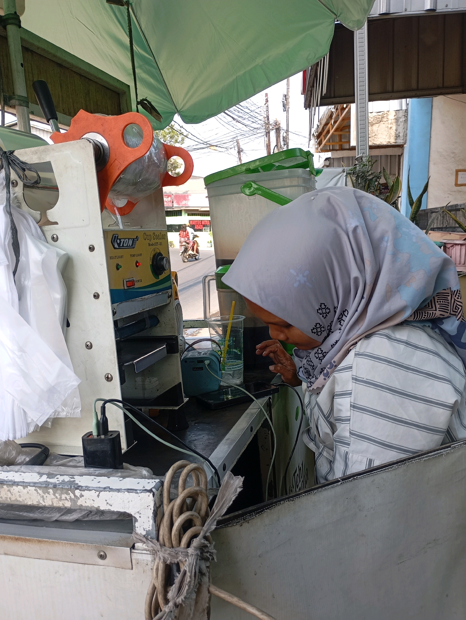 In Babelan, Indonesia, a woman wearing a grey and blue patterned hijab and a striped shirt is operating a cup sealing machine outdoors. The machine, labeled "TON Cup Sealer Model ET-D1," has an orange handle and a yellow control panel with indicator lights for "HEAT LIGHT" and "TEMP LIGHT." A glass with a yellow straw is positioned next to the machine, suggesting it's being used to prepare beverages. The scene appears to be a street-side stall or kiosk, with a green awning overhead. In the background, electrical wires, buildings, and a person on a motorcycle are visible, indicating an urban or semi-urban environment. The overall impression is of a small business operation in a busy street.
