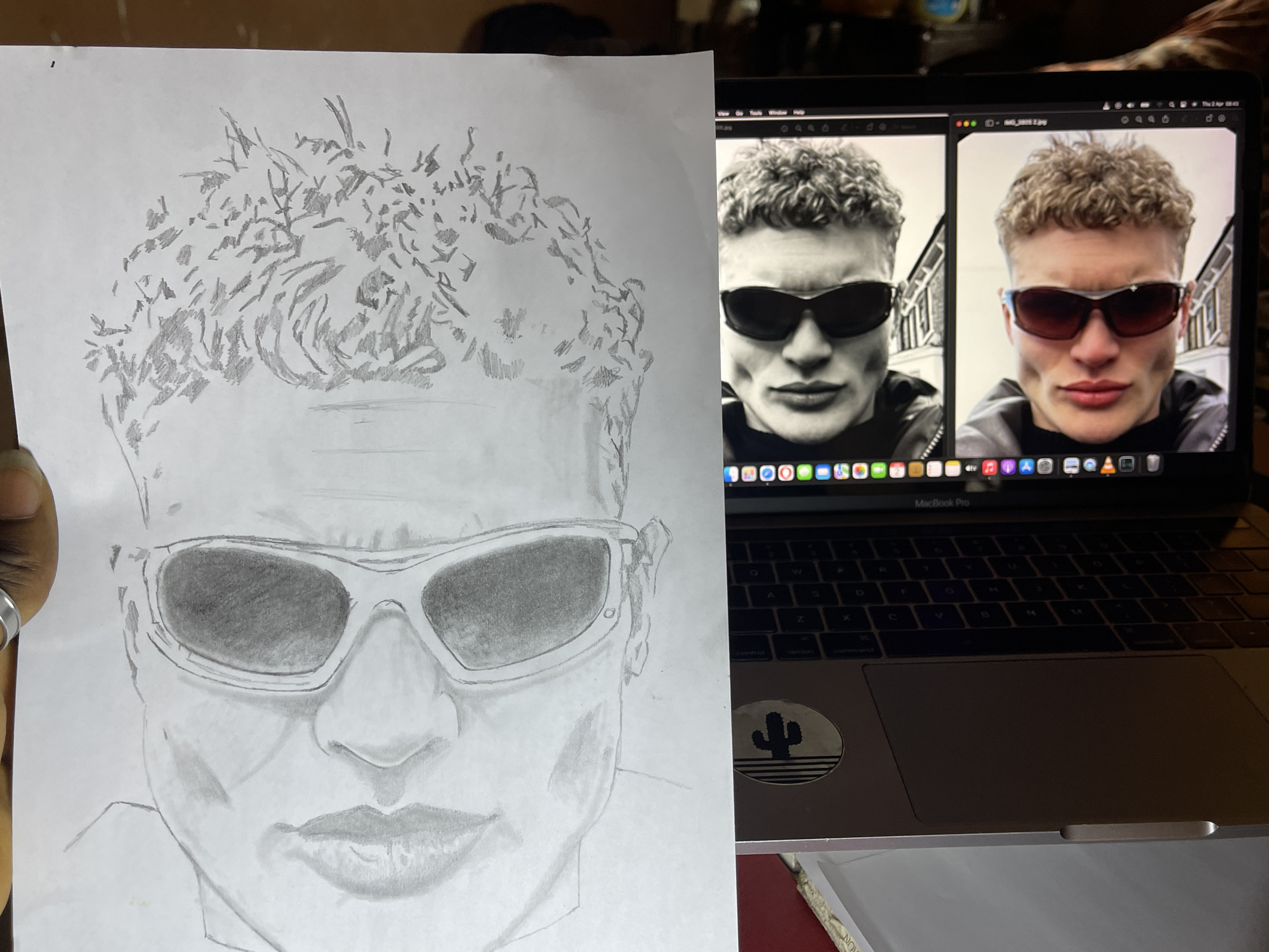The image shows a hand holding a pencil drawing of a person wearing sunglasses, positioned next to a laptop displaying two images of the same person wearing sunglasses. The drawing is on a piece of white paper, showing the person's head and shoulders. The laptop is a MacBook Pro with a black keyboard and a sticker featuring a cactus. The room appears to be indoors and dimly lit. The person in the photographs and the drawing has short, curly hair and a defined jawline.