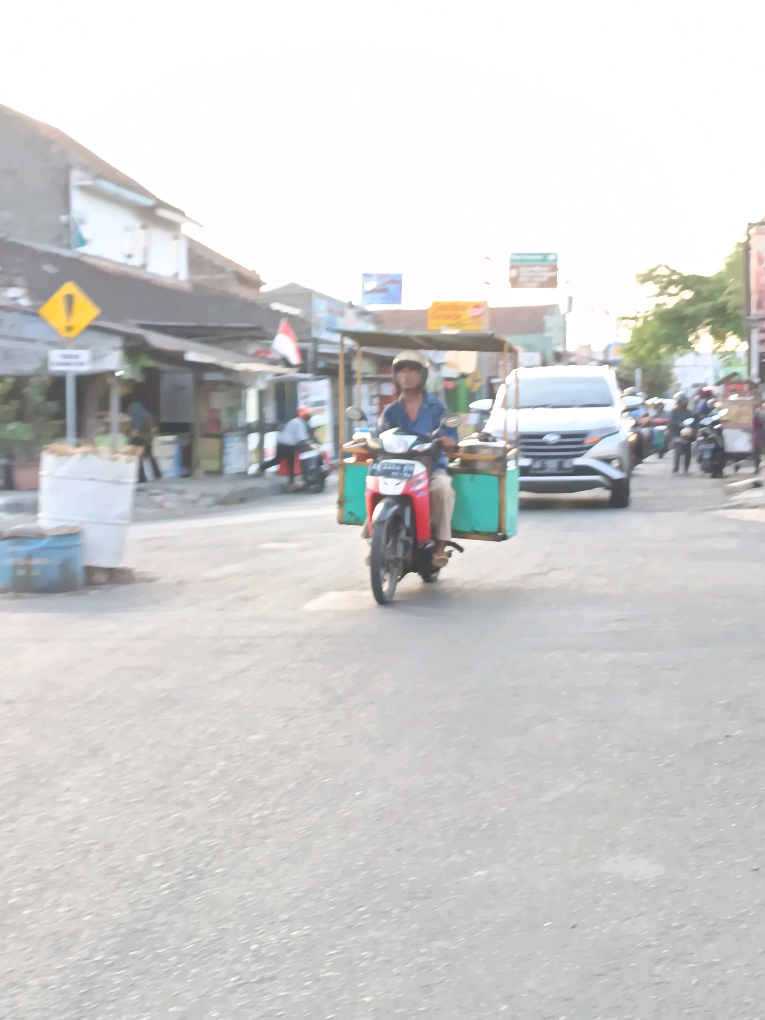 A man is riding a motorcycle with a sidecar in Gatak, Indonesia. The motorcycle is red and white, and the sidecar is green. The man is wearing a blue shirt and a helmet. He is in the middle of the road, and there are other vehicles and buildings in the background. The scene appears to be a street in a village or town, with a mix of residential and commercial buildings. The overall atmosphere is casual and everyday.