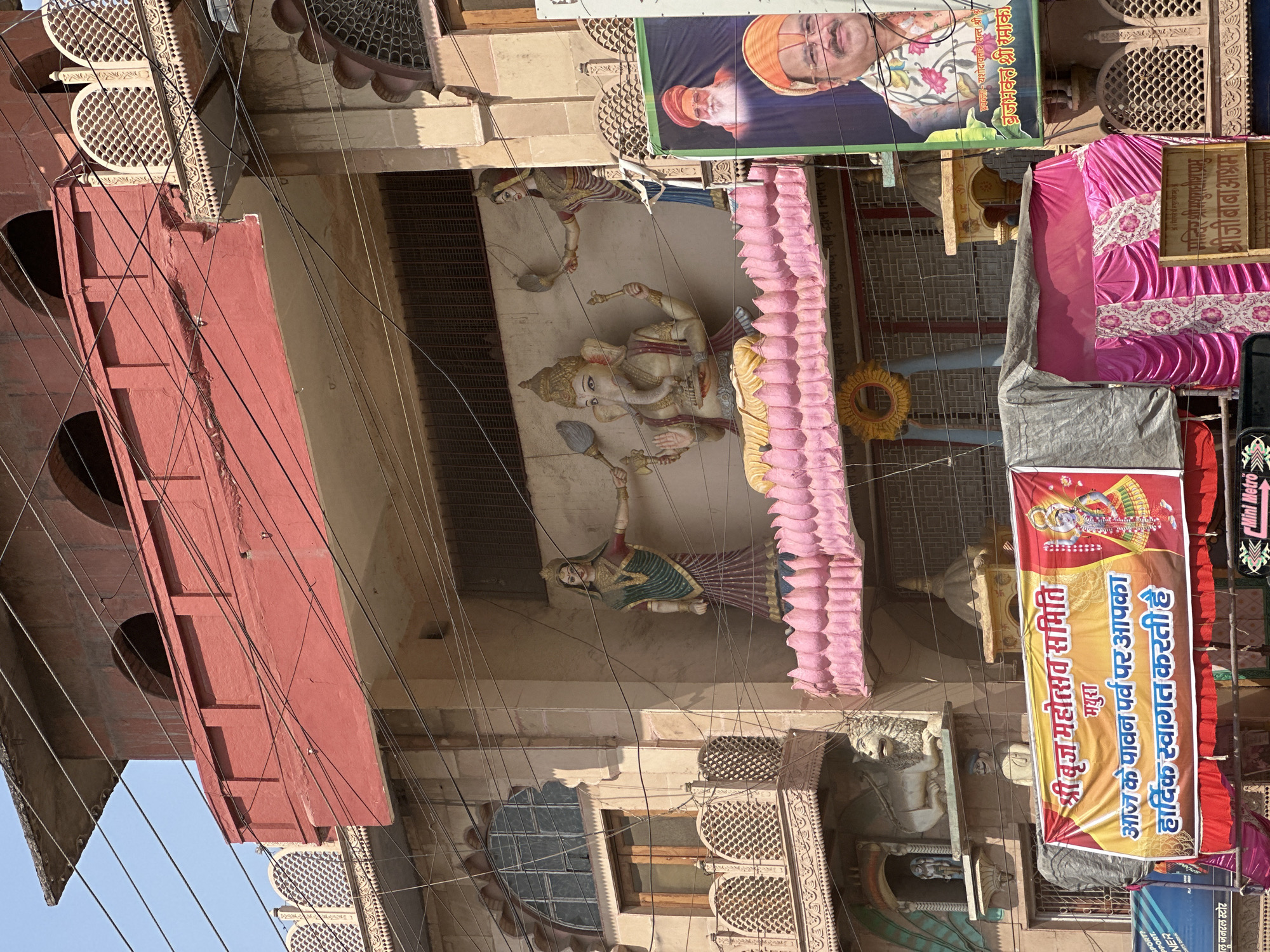 The image depicts a street scene in Mathura, India, likely during a festival or religious event. The perspective is from below, looking up at building facades adorned with decorations, posters, and religious iconography. A prominent banner advertises "Shree Braj Mahotsav Samiti" and welcomes people to a holy festival in Mathura, suggesting a celebration of the region of Braj. Another banner shows a portrait of a man in a saffron turban. Sculptures of deities, including Ganesha, are mounted on the buildings, embellished with pink garlands. The scene is further characterized by an intricate network of electrical wires crisscrossing the sky and the presence of ornate latticework windows. The buildings exhibit diverse architectural styles, some with red facades and arched openings.
