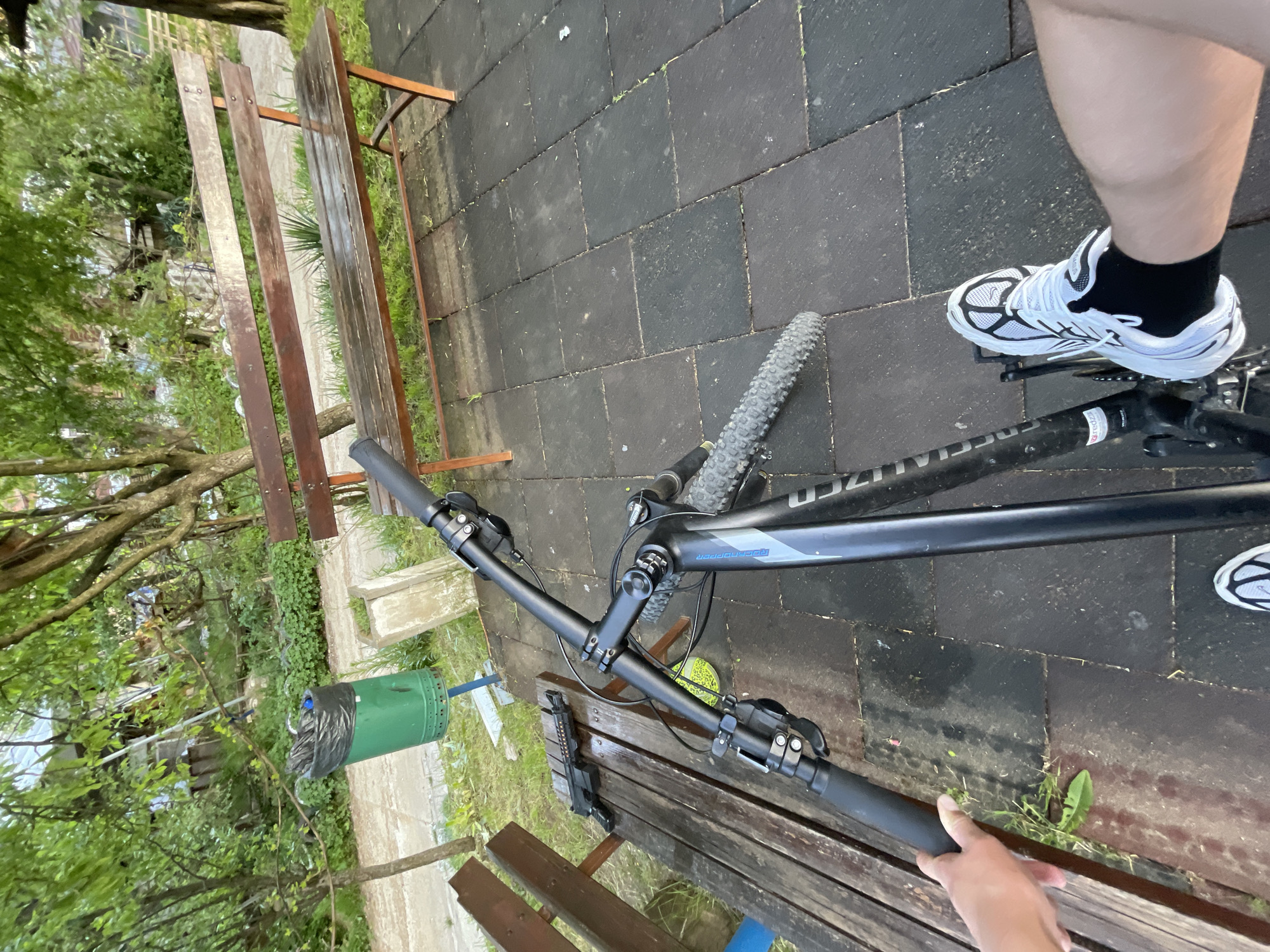 The image depicts a bicycle resting on a wooden bench in an outdoor setting, likely a park or recreational area. The bicycle is dark-colored with a front tire visible, and the handlebars are positioned on the bench. A person wearing white sneakers with black socks is visible standing beside the bike, with their foot on the pedal. The ground is paved with dark gray rectangular blocks. Part of a green trash can is seen in the background near some foliage, including trees and grass. The location is potentially in the city of Zlatograd, Bulgaria, but this cannot be confirmed from the image alone.