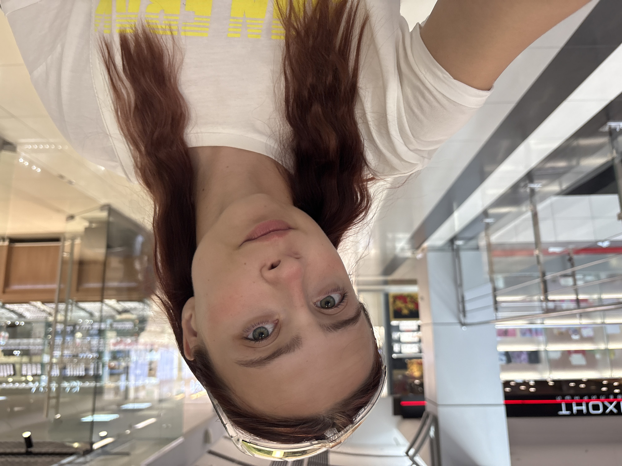 The image is a selfie taken by a young woman with auburn hair and green eyes. She is wearing a white t-shirt and a clear headband with a decorative pattern. The background suggests an indoor location, possibly a shopping mall or a commercial building in Izhevsk, Russia. She is standing in what appears to be a hallway with polished floors and railings. To her left, a sign with red lettering and a red line is partially visible, likely indicating a store or business. To her right, a glass display case filled with items is visible, suggesting a retail environment. The lighting is bright, typical of commercial spaces.