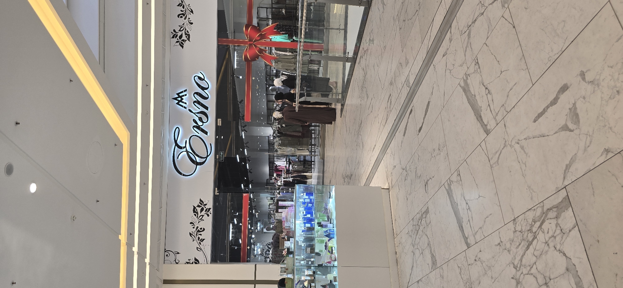 A medium shot captures the facade of the "Ersno" store in Grozny, Russia. The store's sign, featuring stylized script, is prominently displayed above the entrance. A large red bow is seen above the entrance. Inside the store, various items are on display. The foreground shows white marble-patterned flooring. The image is taken at an oblique angle.