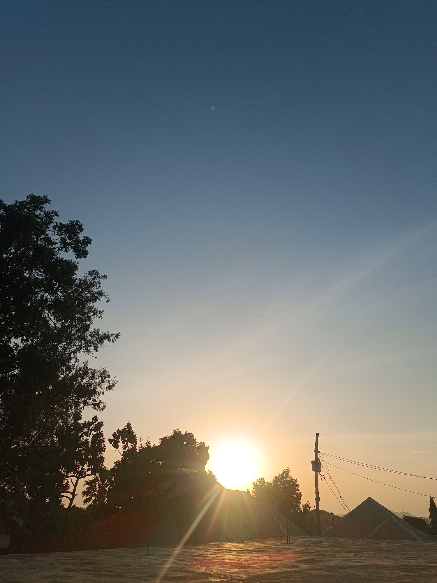 The image depicts a sunrise or sunset scene in Bukuru, Nigeria. The sun is low on the horizon, obscured by trees and buildings, casting a bright glare and sunbeams across the rooftop in the foreground. A silhouetted tree is visible on the left. In the right, there's a utility pole with wires and several low buildings with varied roof types. The sky transitions from a deep blue at the top to a lighter, golden hue near the horizon. The rooftop is flat and appears to be made of concrete. The location is likely a residential area within Bukuru. There are no people visible in the image.