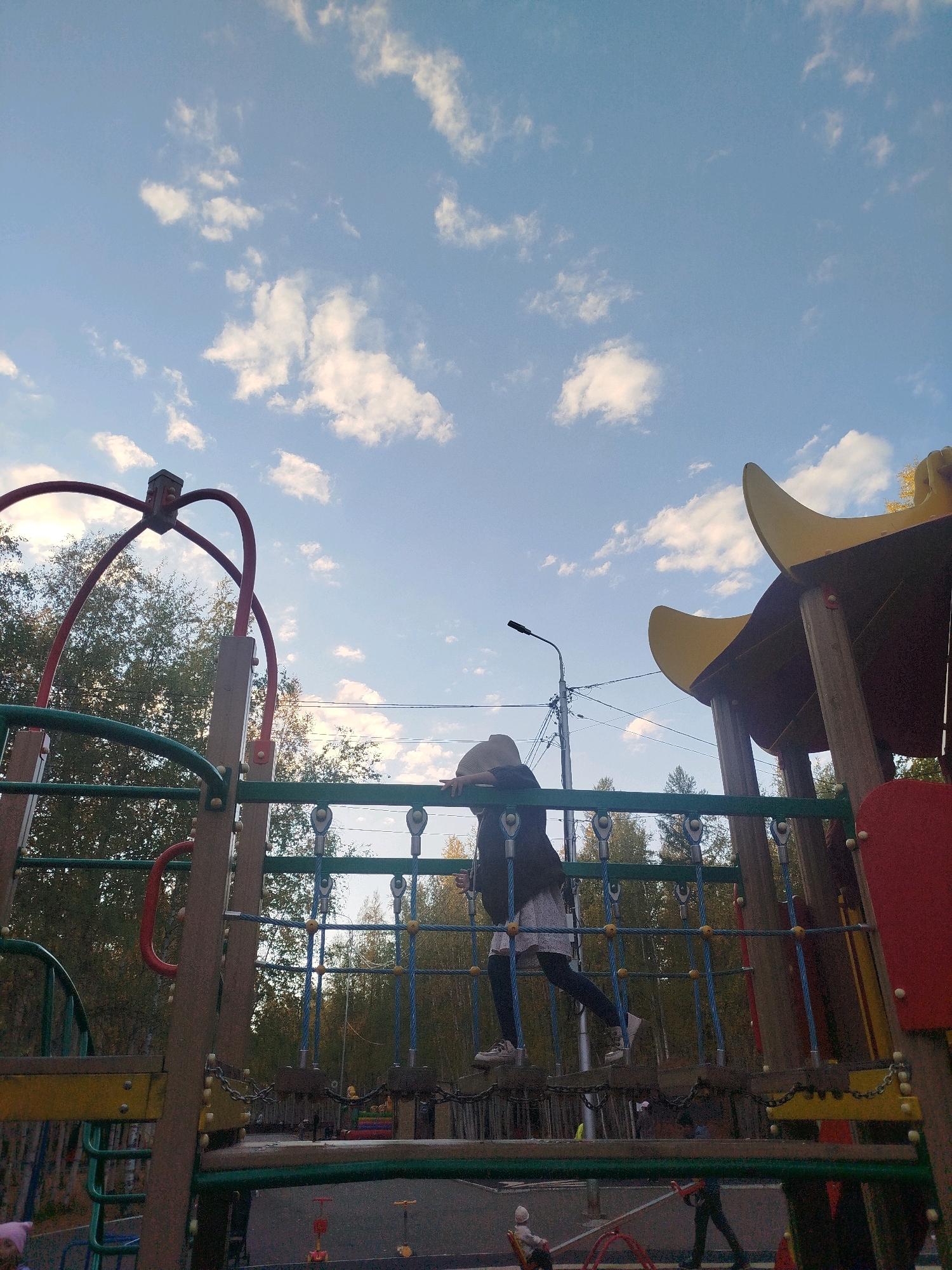 A full shot captures a playground under a partially cloudy blue sky in Mirny, Russia. A young child, wearing a hat and dark clothing over a light-colored dress, is actively navigating a rope bridge on the playground. The playground structure features wooden beams, green and red metal bars, and yellow curved roof elements. Trees with autumn foliage are visible in the background, suggesting the season. A street lamp and utility wires are also present in the scene. In the lower portion of the image, other playground equipment and the ground are visible, with a few other individuals partially obscured.