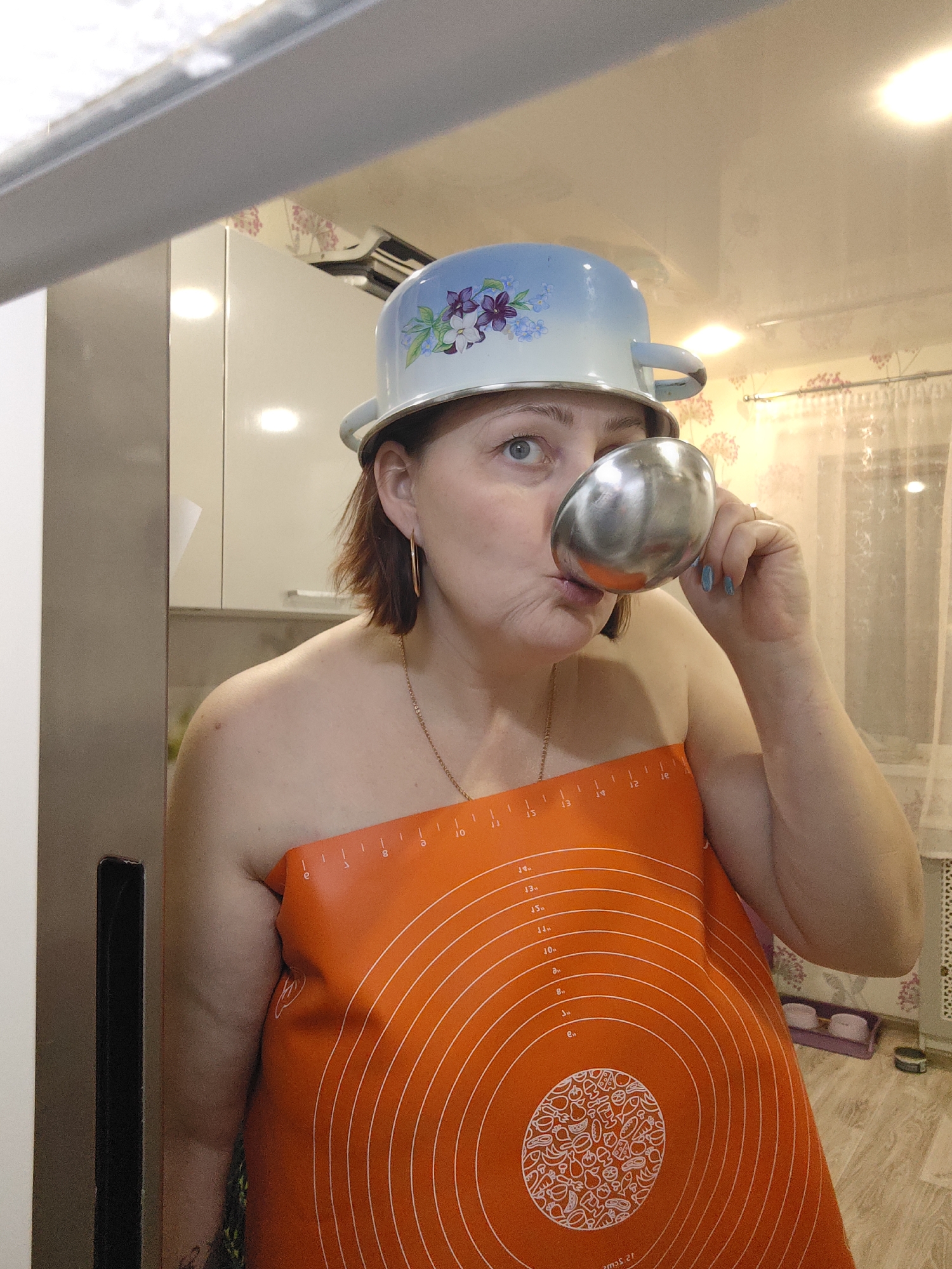 A woman playfully poses in a kitchen, likely in Markovskii, Russia. She is fair-skinned with reddish-brown, medium-length hair and blue eyes, and appears to be middle-aged. On her head, she wears a light blue enameled pot, decorated with purple and green floral patterns, upside down like a hat. She holds a shiny silver ladle or measuring cup to her pursed lips, looking upward and to the right with a wide-eyed, humorous expression. She wears a gold chain necklace and gold hoop earrings, and her fingernails are painted light blue.
Her torso is covered by an orange silicone baking mat, which serves as a makeshift garment. The mat displays concentric circles marked with measurements from 1 to 15 inches and features a central circular design composed of various food item outlines (fruits, vegetables, bread, fish, etc.). Partial text, such as "AMAS.ST" (when viewed upside down from the image perspective), is also visible on the mat.
The background shows a domestic kitchen with light-colored cabinets and countertops. A reflection of ceiling lights and the ceiling itself is prominent in the upper part of the image, possibly from a mirror or highly reflective surface. To the right, a window with sheer white curtains and a wall adorned with pinkish floral wallpaper are visible. The scene conveys a lighthearted and whimsical mood.