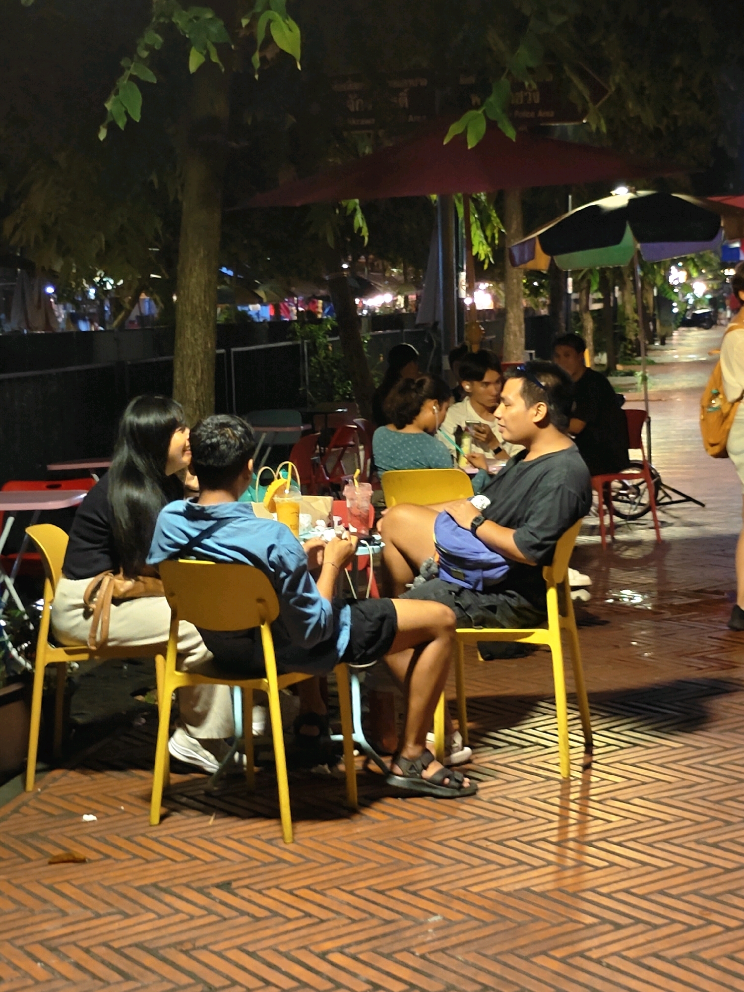 In Bangkok, Thailand, at night, a group of people are seated at an outdoor table, illuminated by ambient lighting. A man in a dark t-shirt and shorts, with a blue bag slung over his shoulder, is seated at the table, looking to his left. Beside him, a woman with long black hair, wearing a black top and light-colored pants, is also seated, looking towards the man. To their left, another woman in a blue patterned top is visible. In the background, other individuals and tables are present, with trees and umbrellas overhead, suggesting a street-side dining or seating area. The ground is paved with a brick-like pattern.