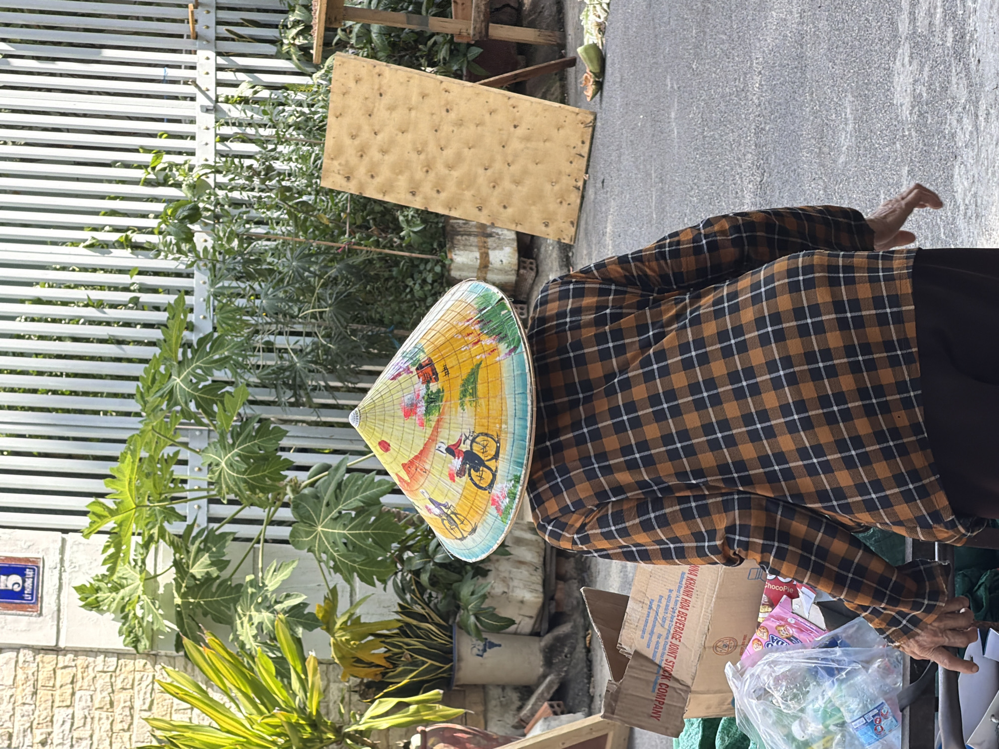 A person wearing a conical hat is standing in front of a white metal fence. The hat is decorated with a painting of people riding bicycles. The person is also wearing a plaid shirt. There are plants and a wooden board in the background. A coconut and some other debris are on the ground. The scene appears to be outdoors, possibly in a market or street setting. The text on the sign in the upper left corner, "5 LY THUONG KIET", indicates the location is Ly Thuong Kiet street, number 5, in Nha Trang, Vietnam.