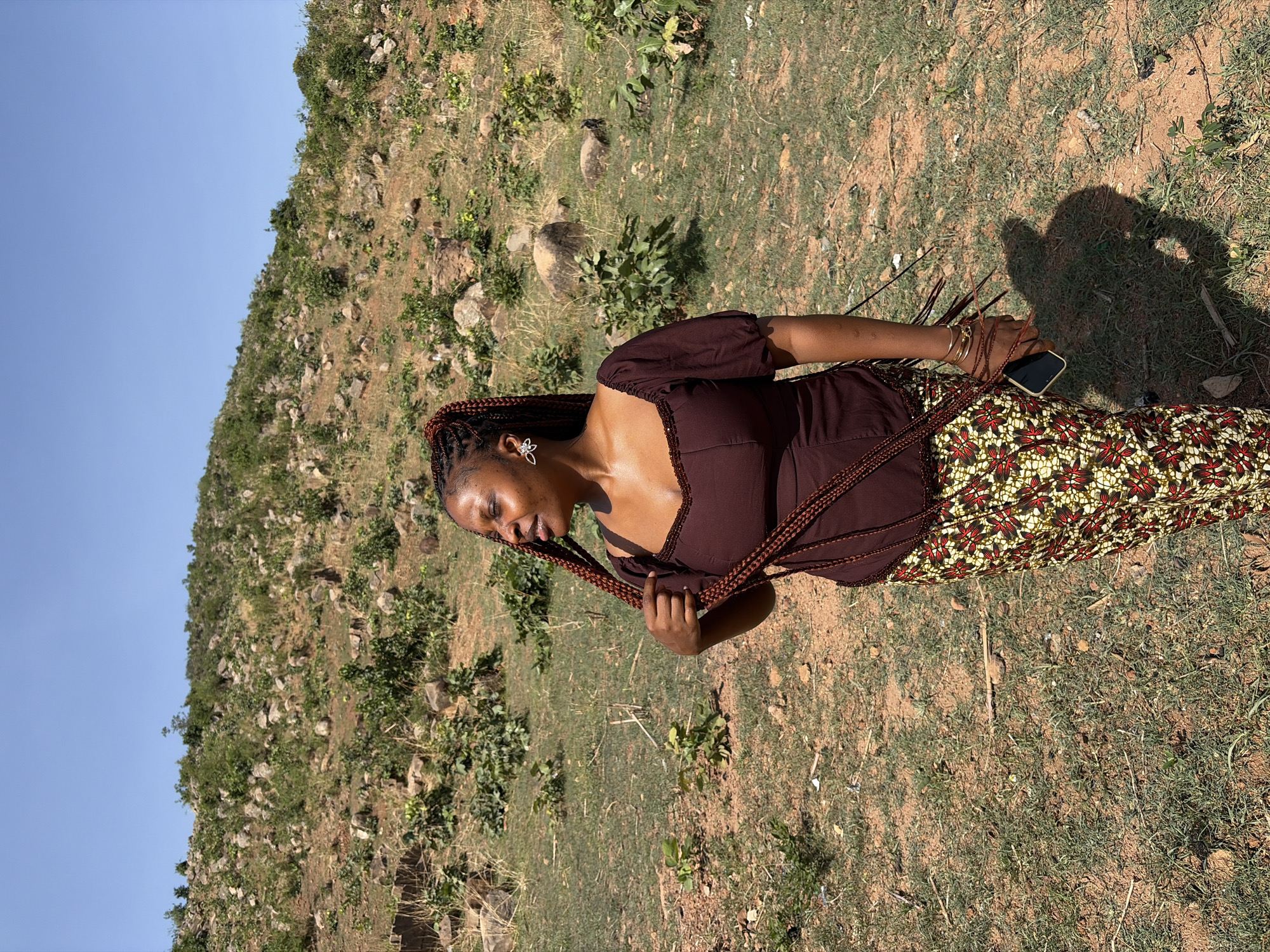 A woman stands in a grassy field with a hillside in the background. The location is potentially near Jos, Nigeria, although this is not explicitly confirmed visually. She is wearing a brown puff-sleeved top and a patterned skirt with red and yellow floral designs. Her hair is styled in braids, and she is holding one of the braids in her left hand. A mobile phone and some gold bracelets adorn her right wrist. The ground is covered with grass and dirt, and the background hillside is dotted with sparse vegetation and rocks. The sky is clear and blue. The photo appears to be taken outdoors in natural daylight.