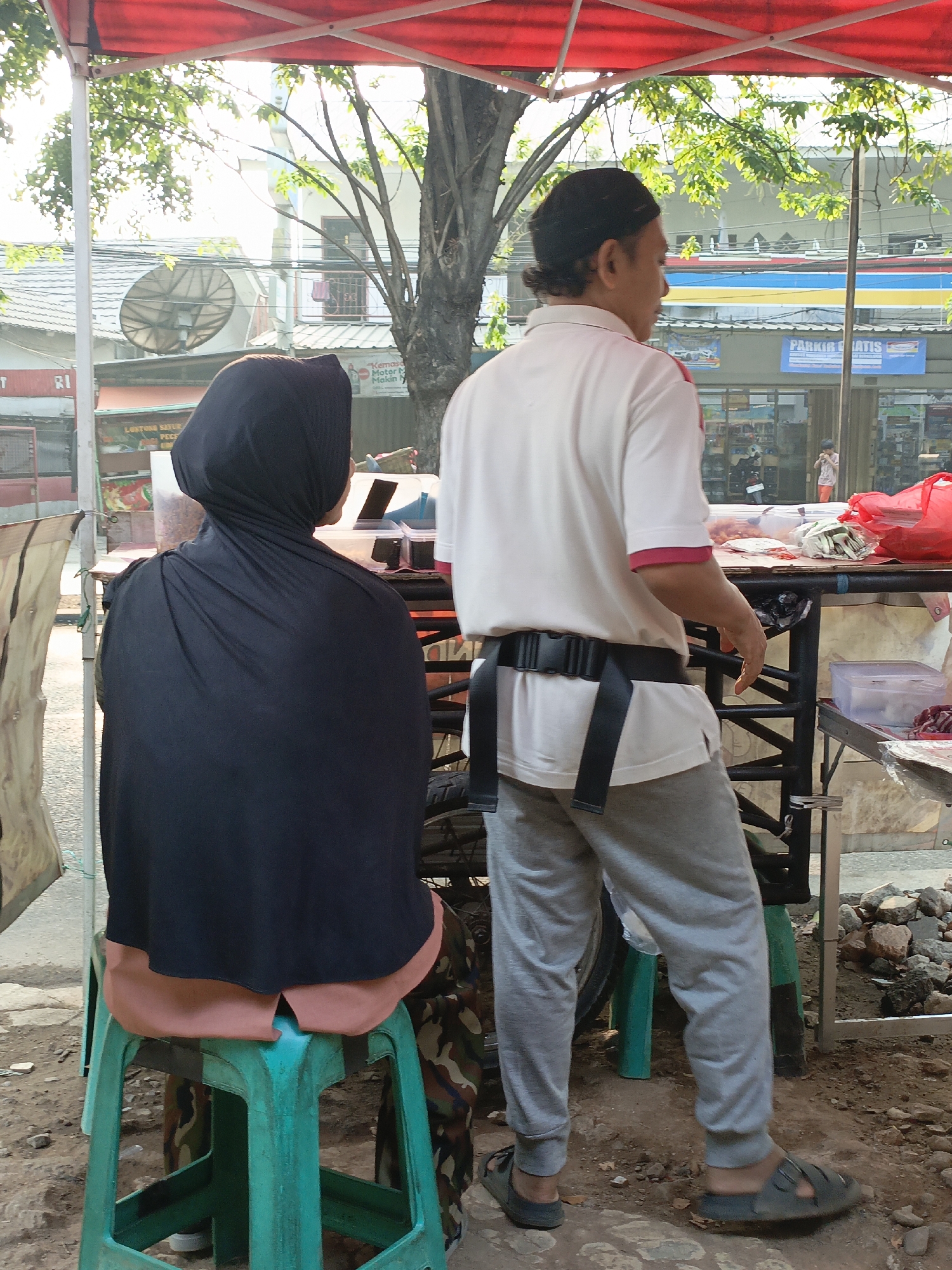 The image depicts two individuals under a red awning in what appears to be a market or street-side stall setting in East Jakarta, Indonesia. On the left, a person wearing a black hijab and a light brown garment is seated on a green plastic stool, facing away from the camera. On the right, a man wearing a white polo shirt with red trim, gray sweatpants, and a black utility belt is standing, also facing away from the camera. He has dark hair pulled back in a bun. Behind them, a black metal table is laden with various items, including red plastic bags, containers, and what looks like food products. The environment shows a street with some rocky ground and a building in the background with visible signage, suggesting commercial activity.