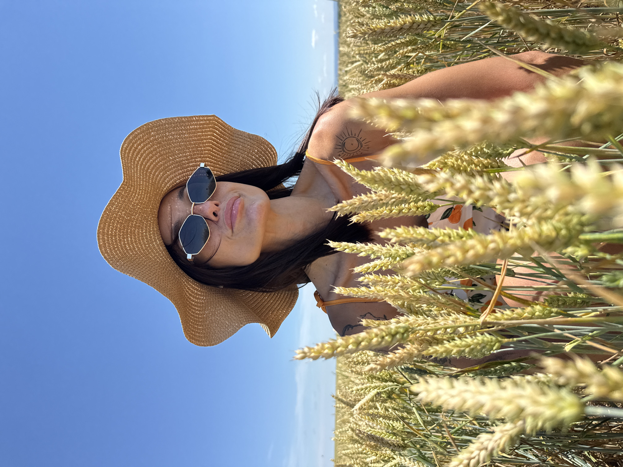 A medium shot captured on a bright, sunny day shows a woman standing in a field of ripe wheat, likely in Zgalevo, Bulgaria. She is wearing a wide-brimmed straw hat and geometric sunglasses, partially obscuring her face as she looks upwards and to her right. Her dark hair is visible beneath the hat, and she has a tattoo of a crescent moon and star on her right shoulder. She is wearing a light-colored dress with an orange floral print and an orange strap visible on her shoulder. The wheat stalks in the foreground are in sharp focus, creating a natural frame for the woman, while the background shows more wheat extending to the horizon under a clear blue sky. The scene evokes a sense of summer warmth and rural serenity.