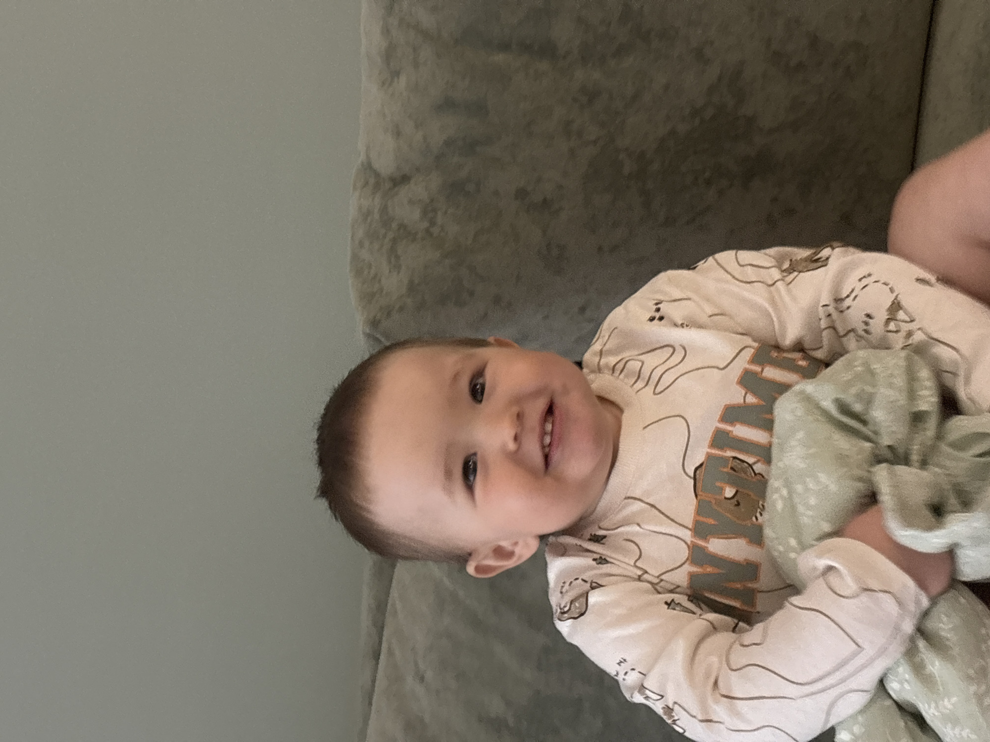 A smiling baby is lying on a gray sofa, holding a light green patterned blanket. The baby has short brown hair and is wearing a long-sleeved shirt with a cartoon design. The background is a light gray wall. The location is unknown and cannot be determined to be Izhevsk, Russia from the given image.