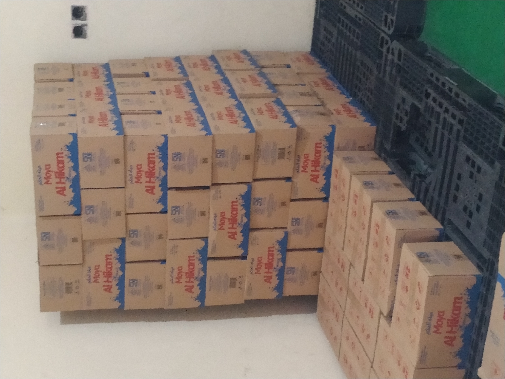 The image shows a tall stack of cardboard boxes labeled "Moya Al Hikam" in what appears to be a storage area. The boxes are brown and have blue lettering and a blue wave design at the bottom. Some of the boxes also have an "SNI" logo. The boxes are stacked on top of black plastic pallets. In the background, there is a white wall with two black electrical outlets. The scene is lit by artificial lighting, and the overall impression is one of a warehouse or storage facility. The location in Batan, Indonesia, is a city known for its industrial areas, which would be consistent with the scene depicted. There are no people visible in the image.