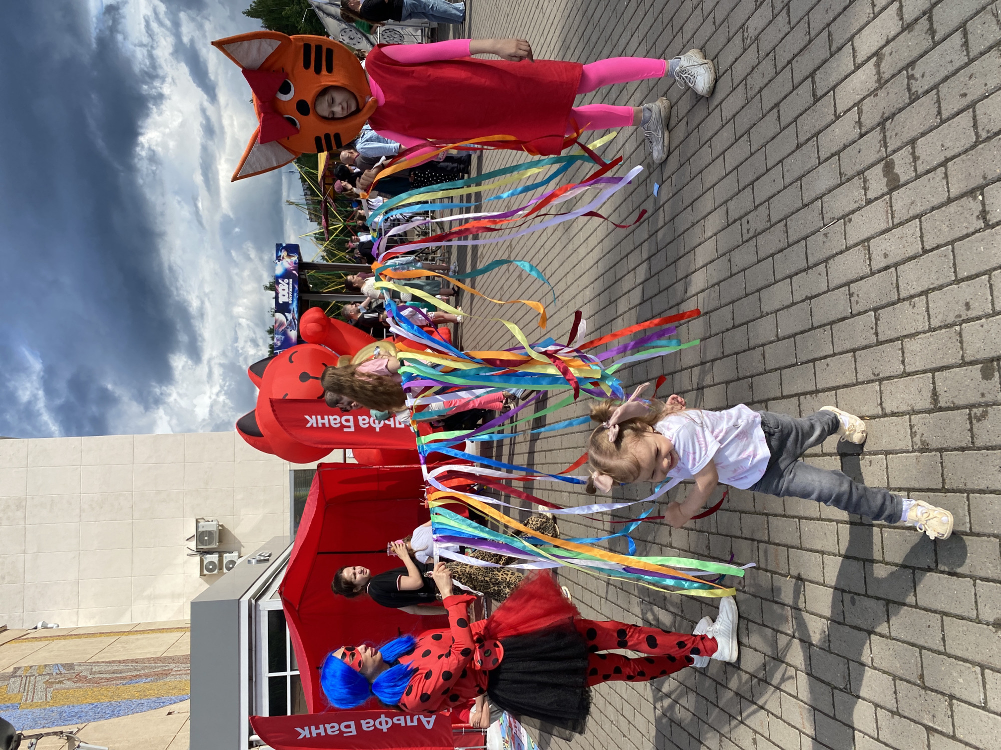 The image depicts an outdoor event, likely a festival or celebration, taking place on a paved area in front of a building. A young girl in a white shirt and gray jeans is running towards the right, holding a string of colorful ribbons. To her left, a person in a Ladybug costume with a blue wig and black and red polka dots is holding the ribbons. To the right of the girl, a person in a cat costume, featuring an orange head with a red bow and pink leggings, also holds the ribbons. In the background, there are large inflatable red cat figures, a red tent, and various people observing the event. Two red banners with white text, "Альфа Банк," are visible on the left and right sides of the frame, indicating a sponsor or location associated with Alfa-Bank. The sky is cloudy, suggesting overcast weather. The scene appears to be on the territory of the Chaikovsky Palace of Culture, located in the city of Chaikovsky, Russia, given the presence of the Alfa-Bank banners, which often sponsor local events and the visible architecture.