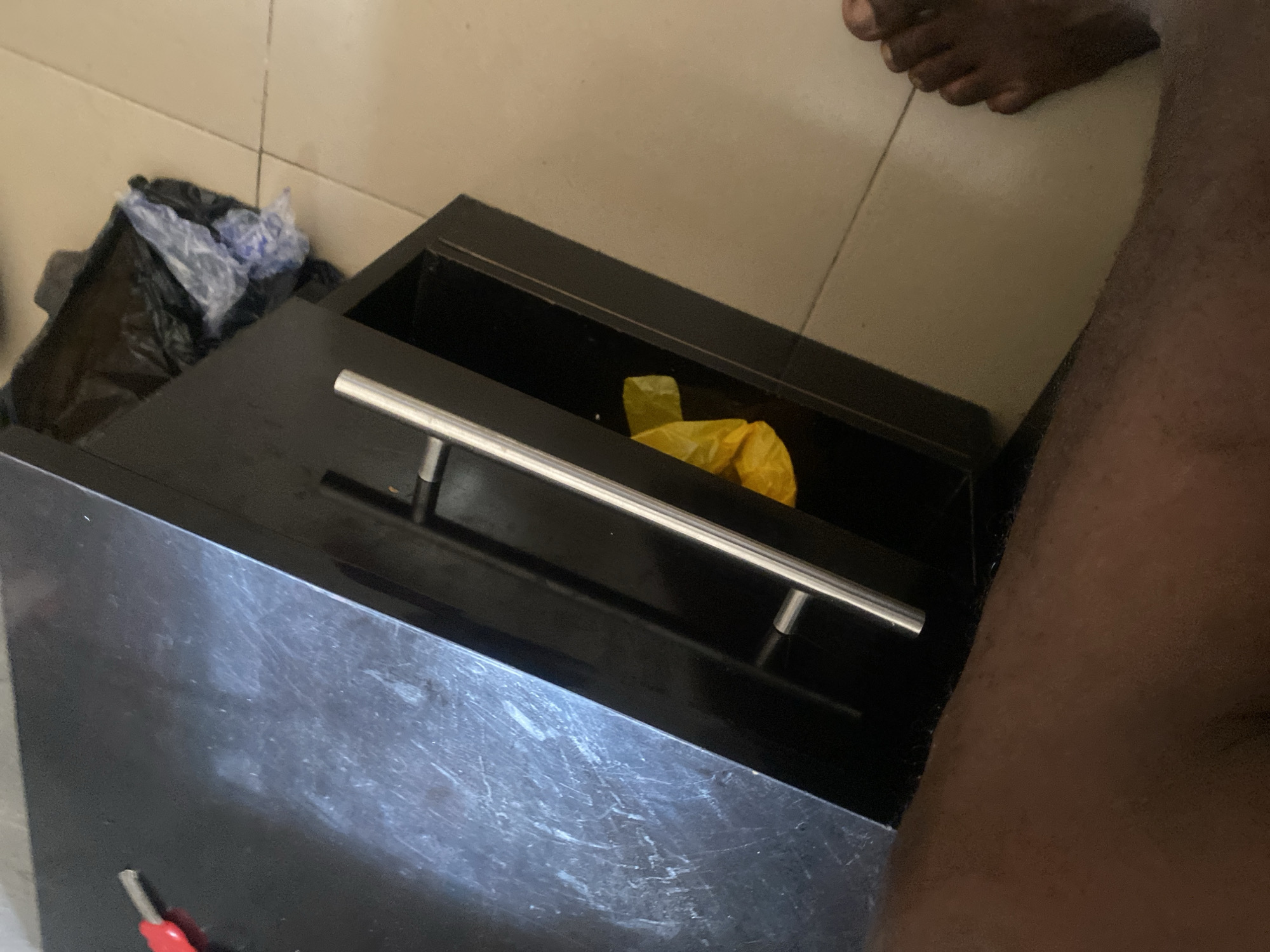 The image depicts a close-up view of a black cabinet with a partially open drawer. Inside the drawer, a crumpled yellow plastic bag is visible. To the left of the cabinet, a red and silver object, possibly a tool, rests on the cabinet's surface. To the right of the cabinet, a person's foot is visible on a tiled floor. In the background, a black trash bag containing crumpled plastic is situated on the floor next to the cabinet. The scene appears to be indoors.