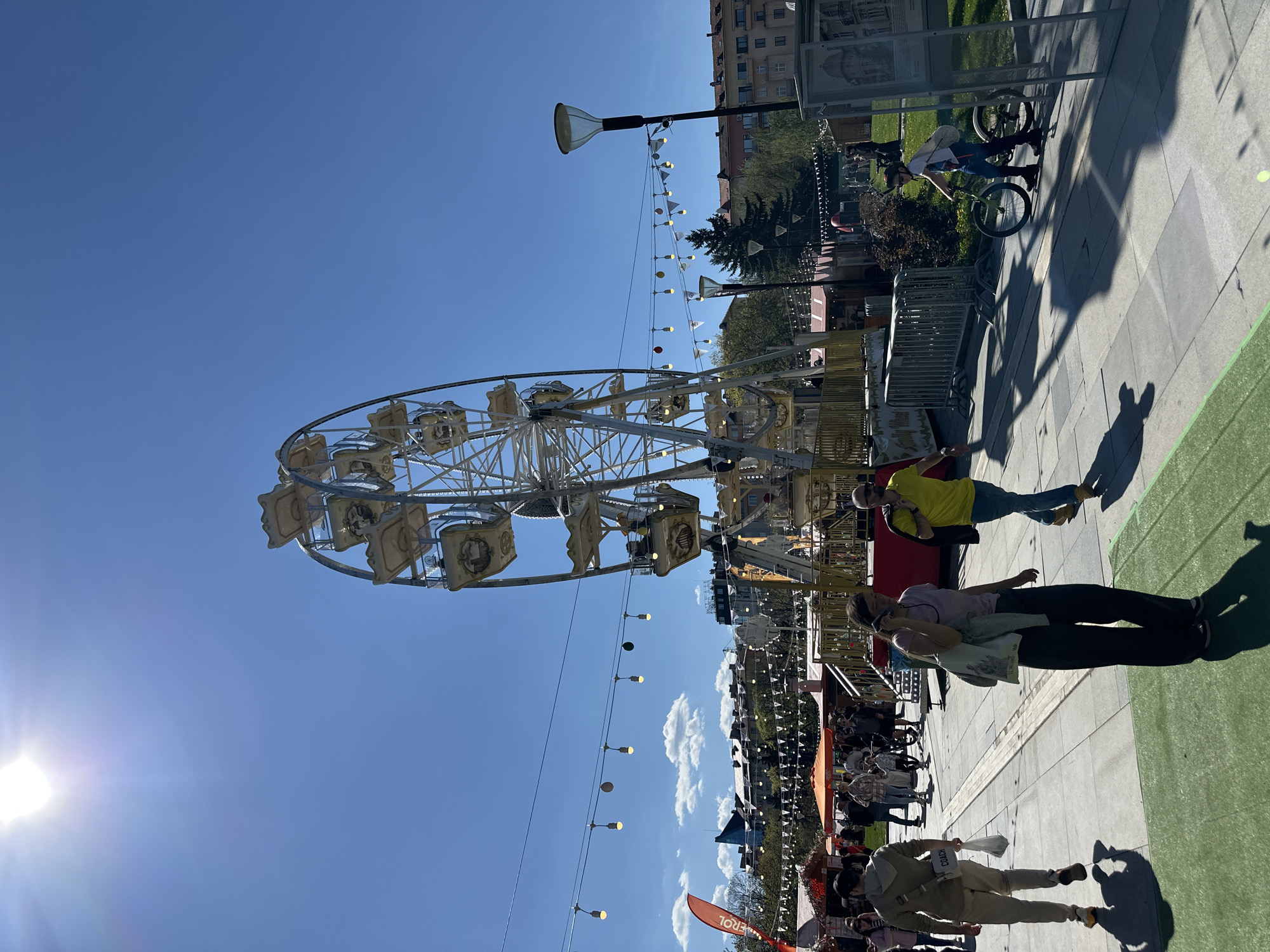 The image shows a sunny day in a public space, likely in the city of Sofia, Bulgaria. A large Ferris wheel is the central focus, under a clear blue sky. Strings of lights are strung around the wheel. There are buildings and trees in the background. A number of people are visible walking or standing, some wearing face masks. One person is riding a bicycle. The ground is paved with concrete and some artificial turf. A display is behind the people with the bicycle.
