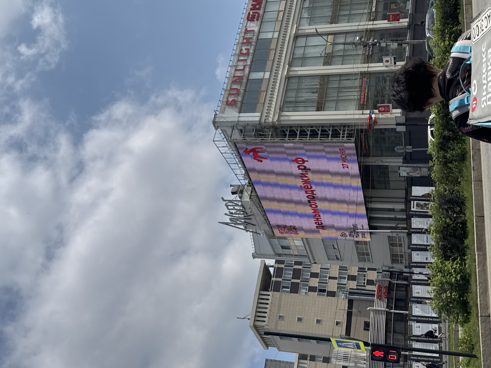This is a photograph of a street scene in Perm, Russia, with a focus on a large digital billboard displaying an advertisement for "деньмолодежи.рф" (Day of Youth) and the date "27 ИЮНЯ" (June 27). The billboard is mounted on the side of a building that also has a large illuminated sign reading "SUNLIGHT" and another partially obscured logo. To the left of the digital billboard, a sign with Cyrillic script reads "АЛЬБЕРТ" (Albert). In the foreground, the back of a person wearing a backpack with "Яндекс Лавка" (Yandex Lavka) branding is visible. A pedestrian traffic signal shows a red walking man, with the number "01" displayed below it. Several other buildings, including a tall, modern structure with a checkered facade, line the street. Green foliage forms a hedge along the sidewalk. The sky is partly cloudy.