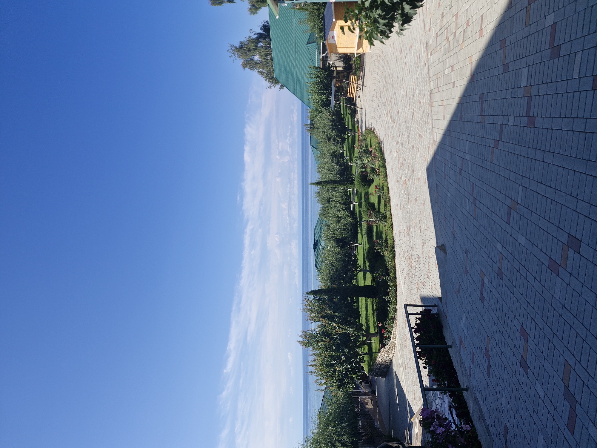 The image depicts an outdoor scene in Bulan-Segettu, Kyrgyzstan, characterized by a paved courtyard leading towards a body of water. The foreground is dominated by a gray and reddish-brown tiled courtyard, with a long shadow cast from an unseen object on the right side. To the left, a black metal railing runs along the edge of the courtyard, bordering a garden area with pink and purple flowers and dark green bushes. Beyond the railing, a stone retaining wall curves away, separating the courtyard from a lush green lawn dotted with trees. In the mid-ground, several trees with green foliage are planted in rows. In the background, a calm, light blue body of water stretches horizontally, meeting a clear blue sky with sparse white clouds. Several buildings with green tiled roofs are visible amongst the trees. One building on the right appears to be a wooden structure with a yellow facade. A wooden swing set is positioned on the lawn near the buildings. The overall impression is of a resort or recreational area by the water on a sunny day.