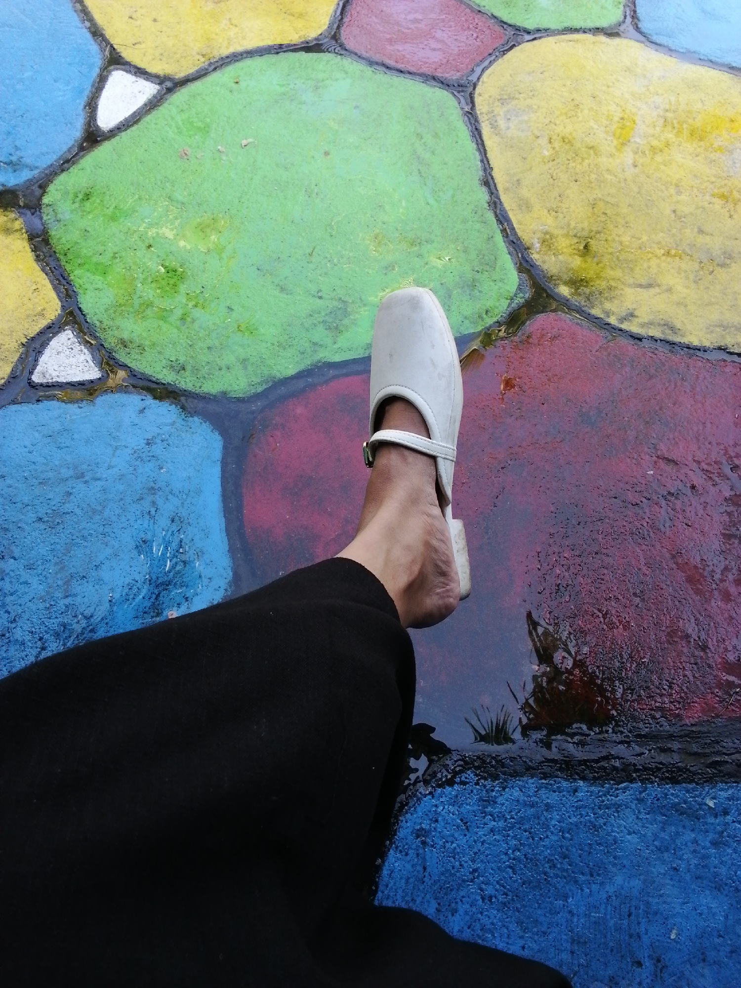 A person's left foot wearing a white sandal and black pants stands on a wet, colorful ground made of large, irregularly shaped stones in Tegalwangi, Indonesia. The stones are painted in various bright colors, including blue, green, yellow, and red, with black grout lines separating them. The ground appears to be outdoors, possibly in a public space, as indicated by the patterned pavement and the wet surface suggesting recent rain or a water feature.
