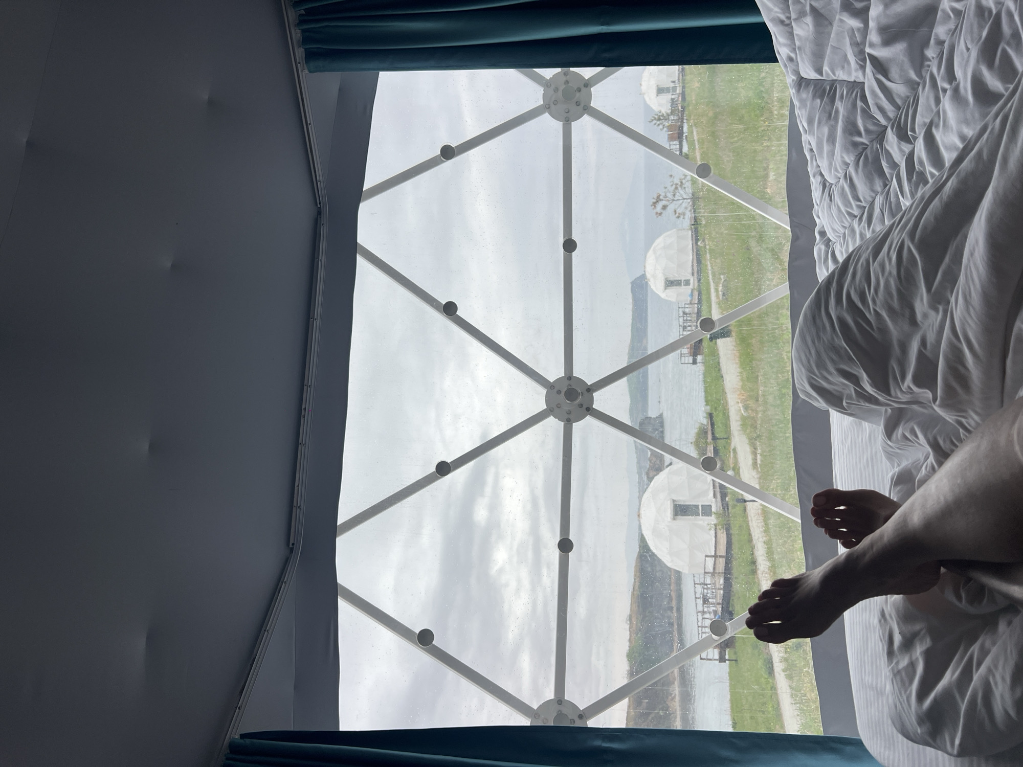 From a bed in what appears to be a geodesic dome in Zarubino, Russia, there is a view of two similar domes on stilts over the water. The weather is overcast. A pair of bare feet are visible in the bed.